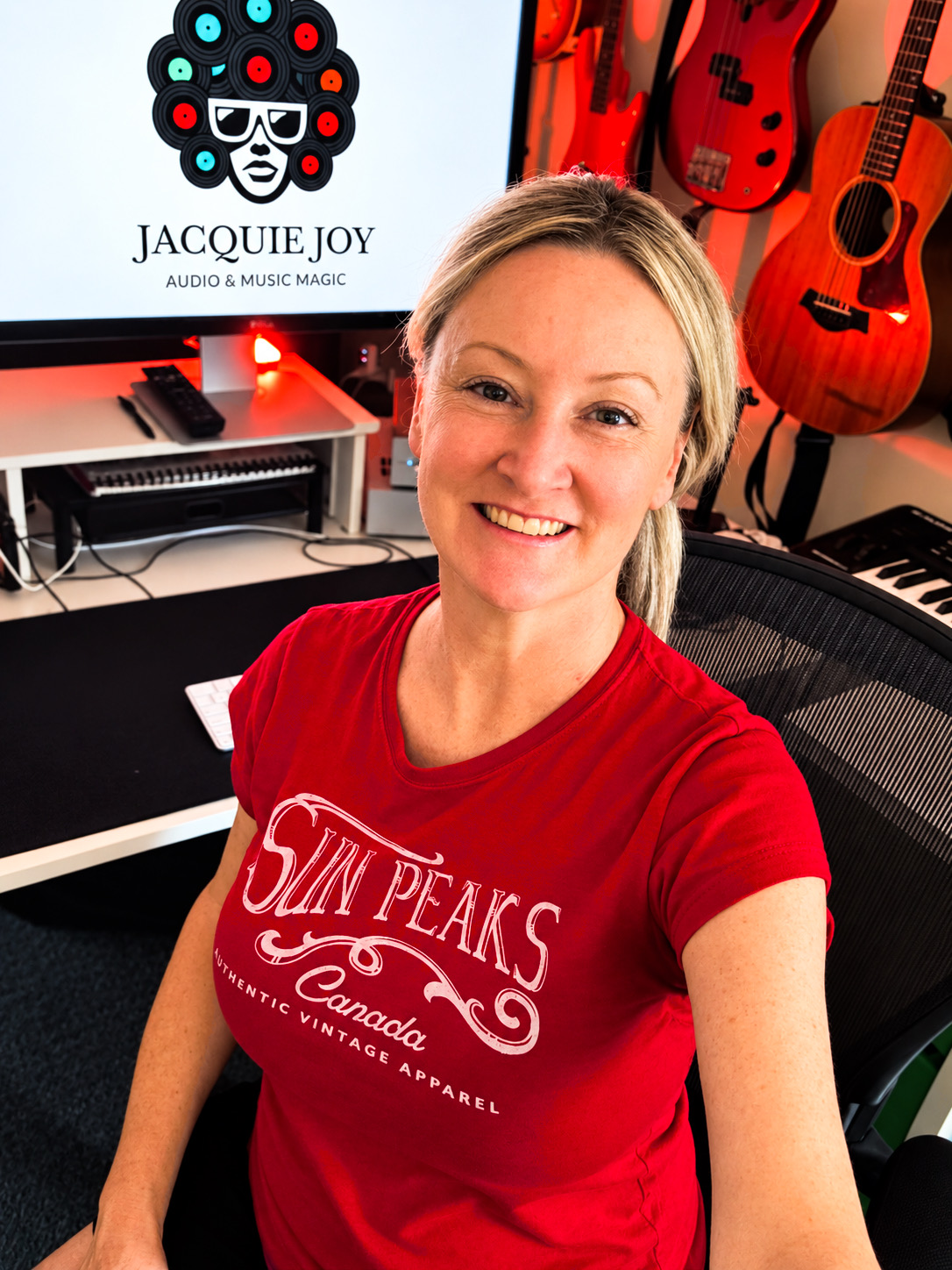Woman with blonde hair smiling in music studio with guitars and keyboards, wearing a red T-shirt with white text.