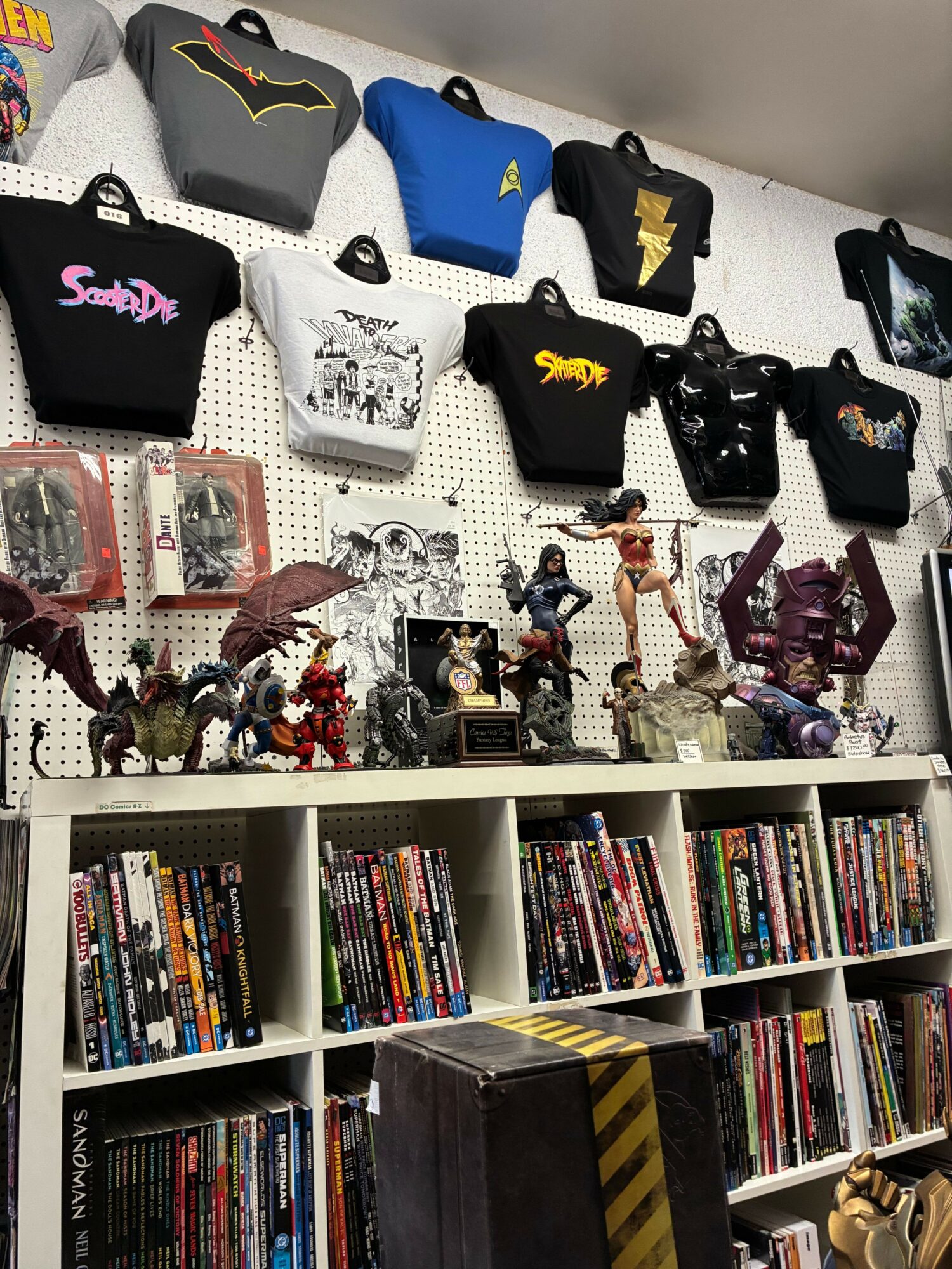 Table with various handbags, jewelry, and figurines, next to shelves filled with DVDs or books, in a store or library.
