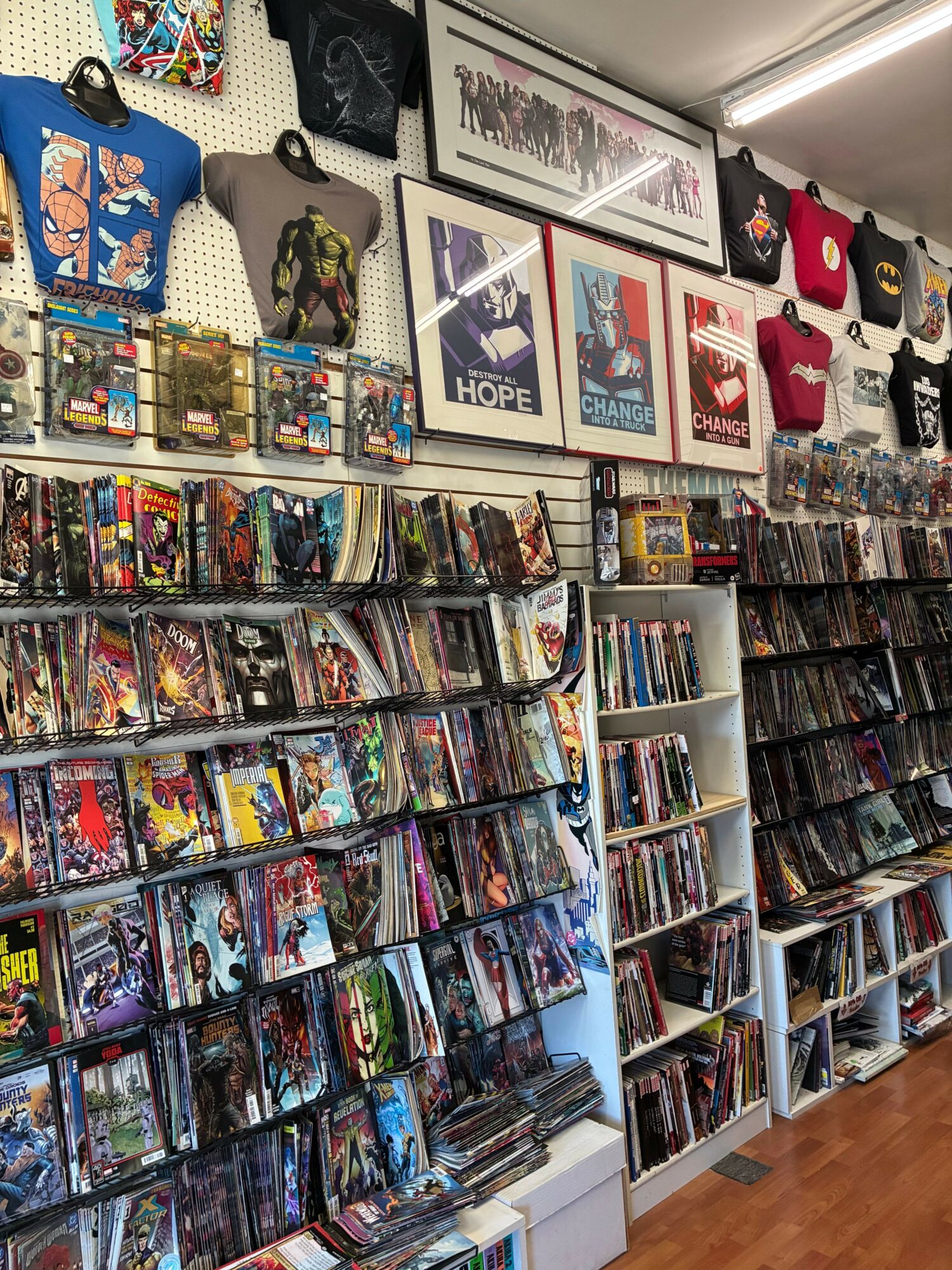 Multiple comic books and graphic novels arranged on tables, with some framed artwork and collectibles visible in the background.