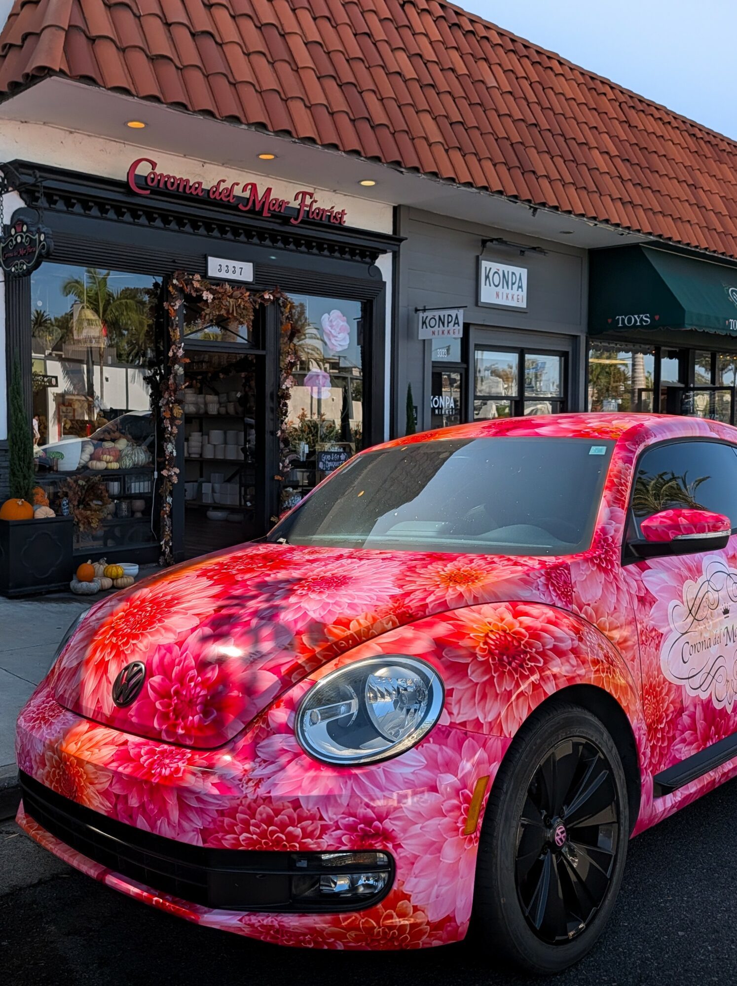 Pink car with floral design parked in front of a storefront with a red-tiled roof.