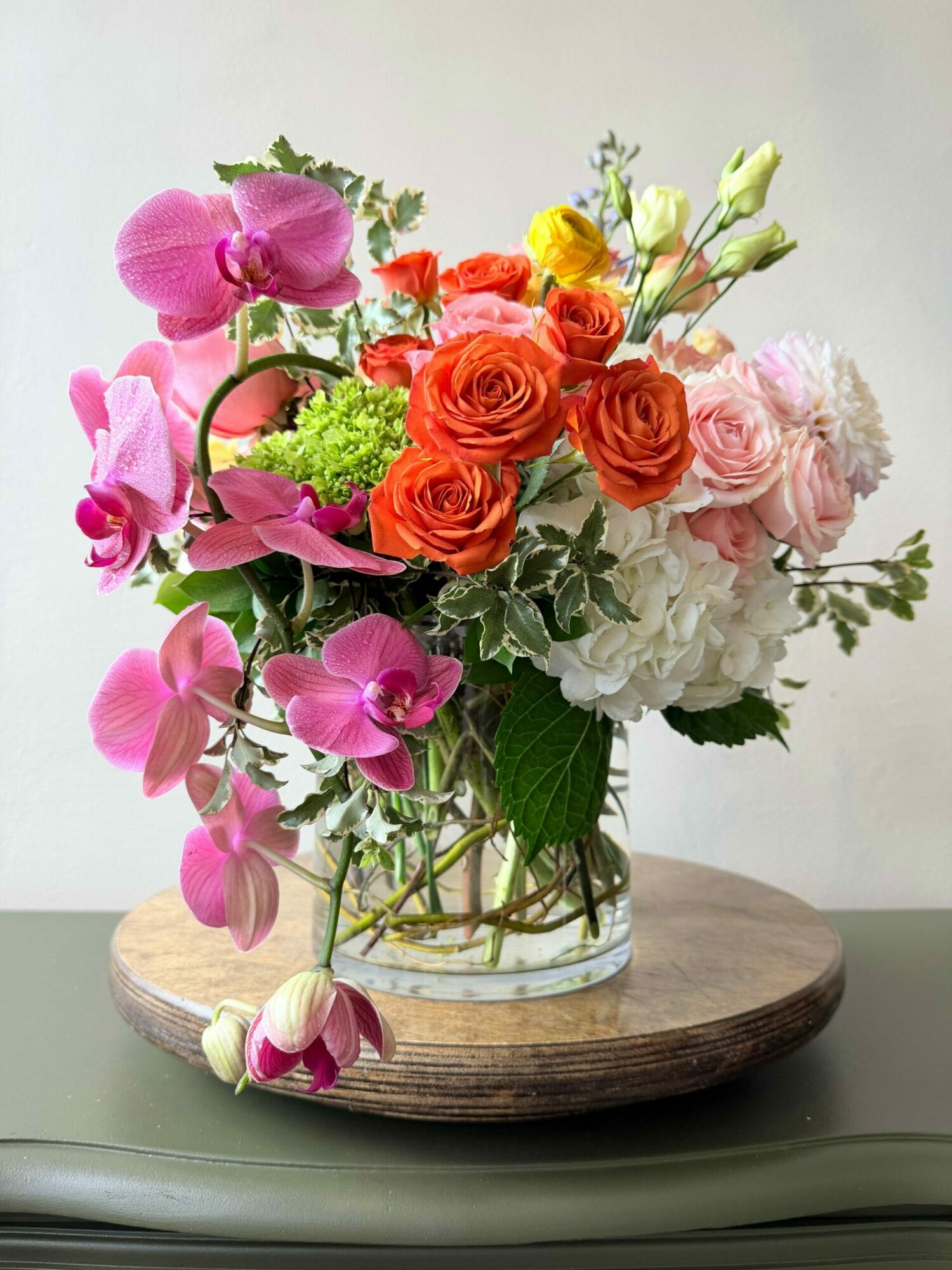 Colorful flower arrangement with roses, orchids, and greenery in a glass vase on a wooden base.