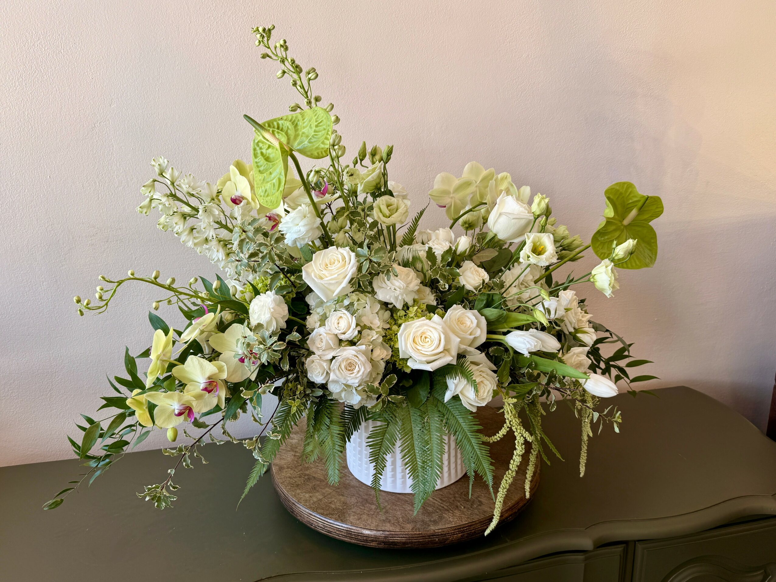 White floral arrangement with roses and greenery in a round wooden container on a dark surface.
