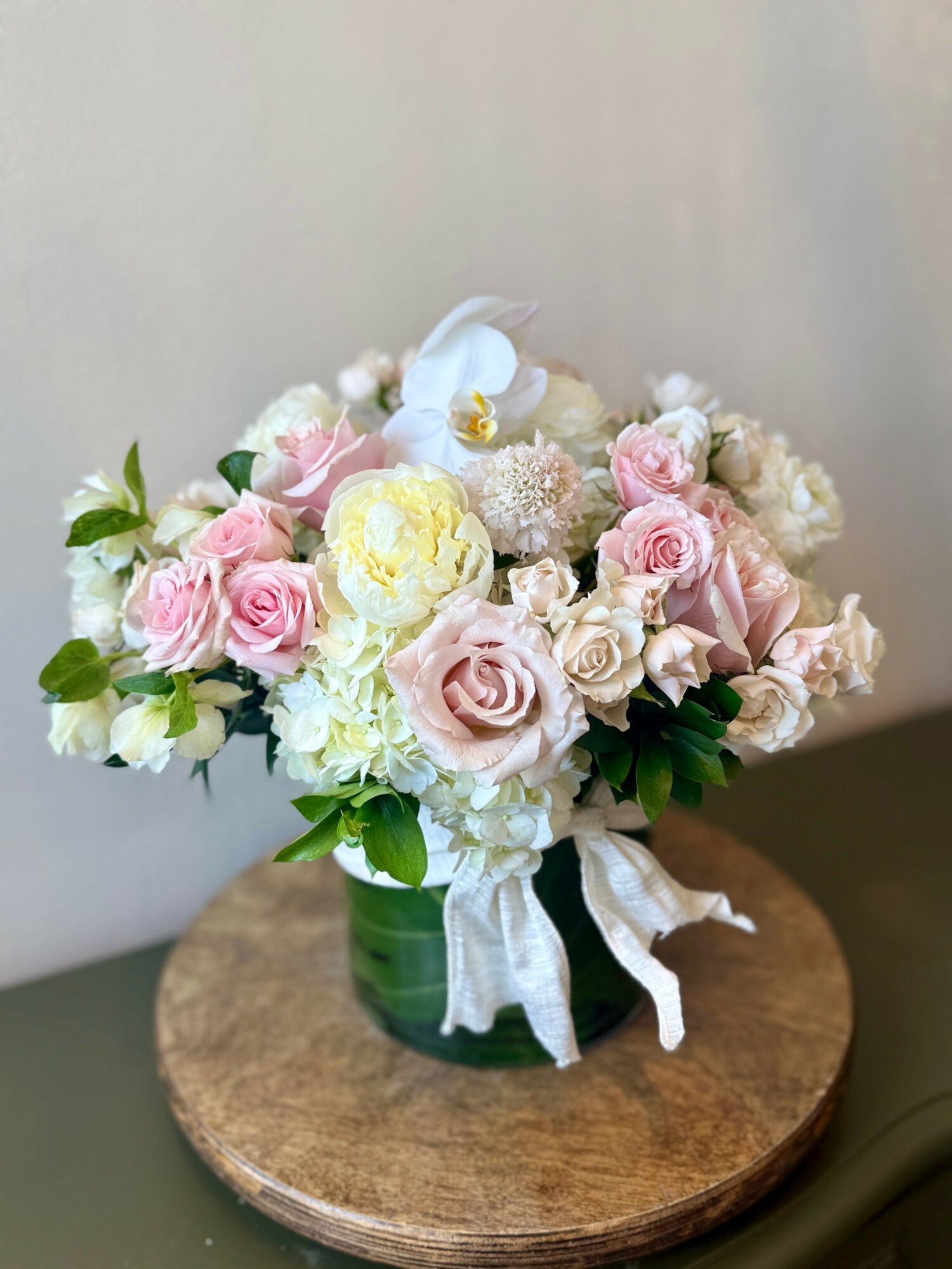 Flower arrangement with pink and white roses, a white orchid, and greenery in a green vase with a white ribbon, on a wooden surface.