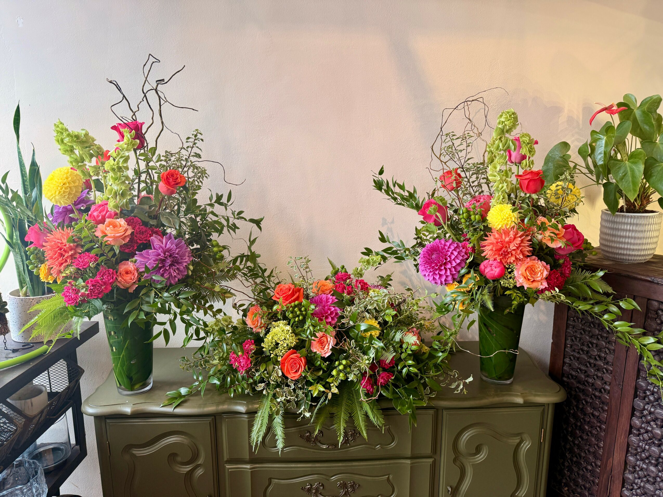 Three colorful flower arrangements in vases on a table, with a potted plant nearby, against a plain wall.
