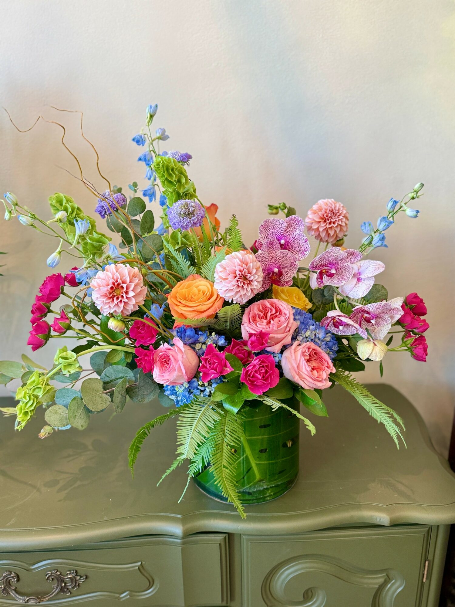 Colorful flower arrangement in a green vase with pink, purple, orange, and blue flowers, and green foliage, on a table.