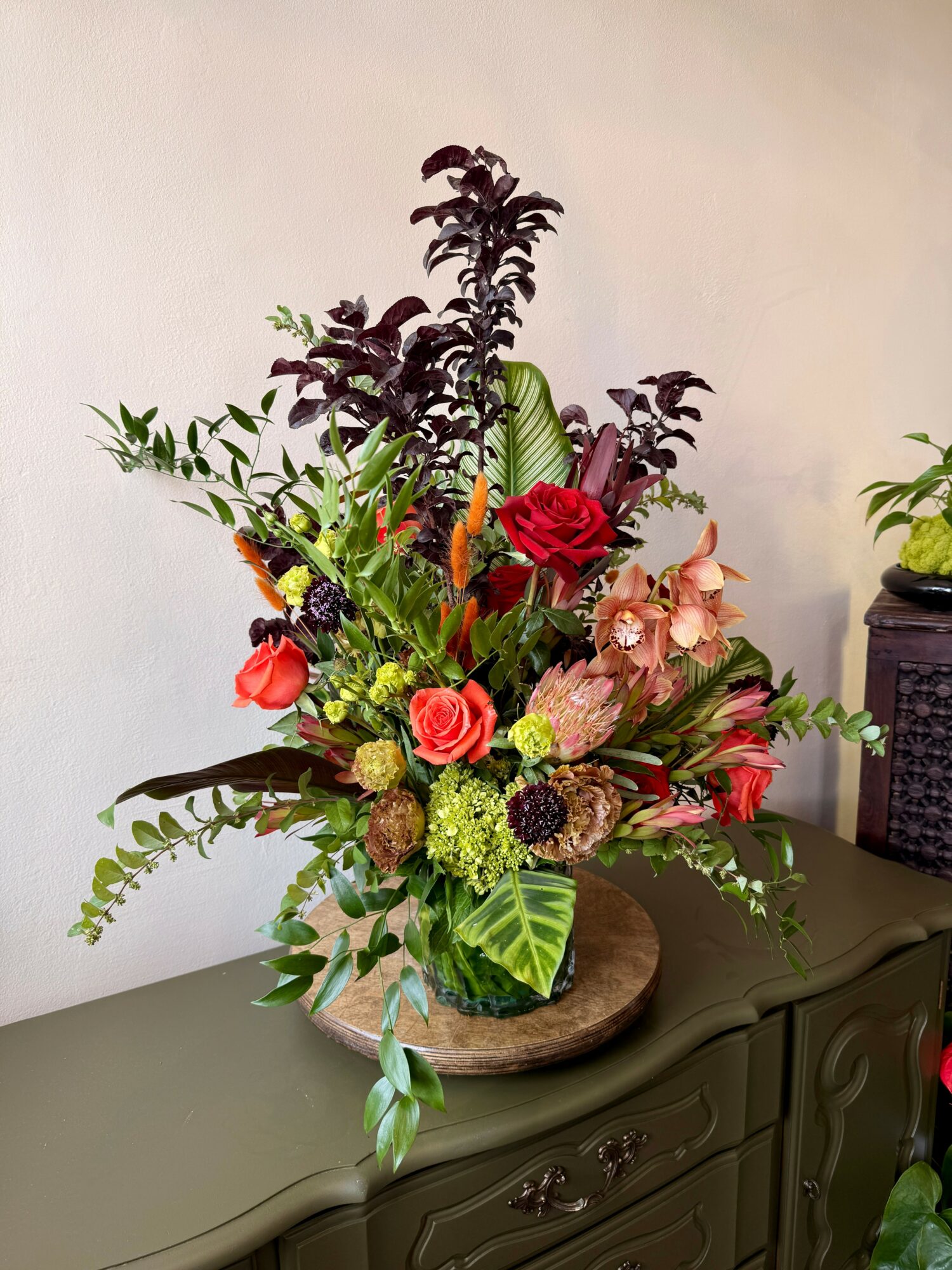 Colorful flower arrangement with roses, greenery, and dark purple leaves in a round container on a dark surface.
