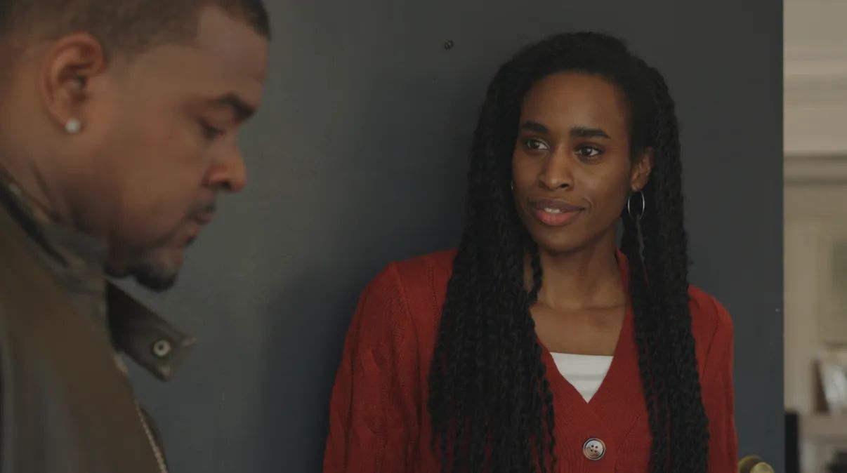 Two people standing indoors, one with a serious expression, the other with a neutral expression, against a dark wall.