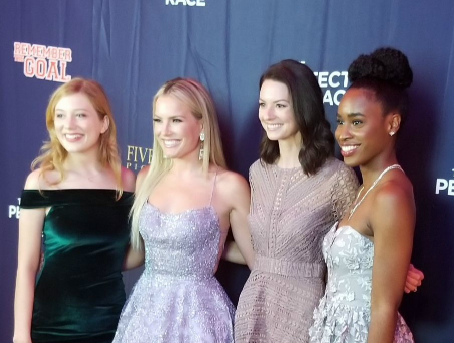 Four women in elegant dresses standing together, smiling, against a blue backdrop with event logos.