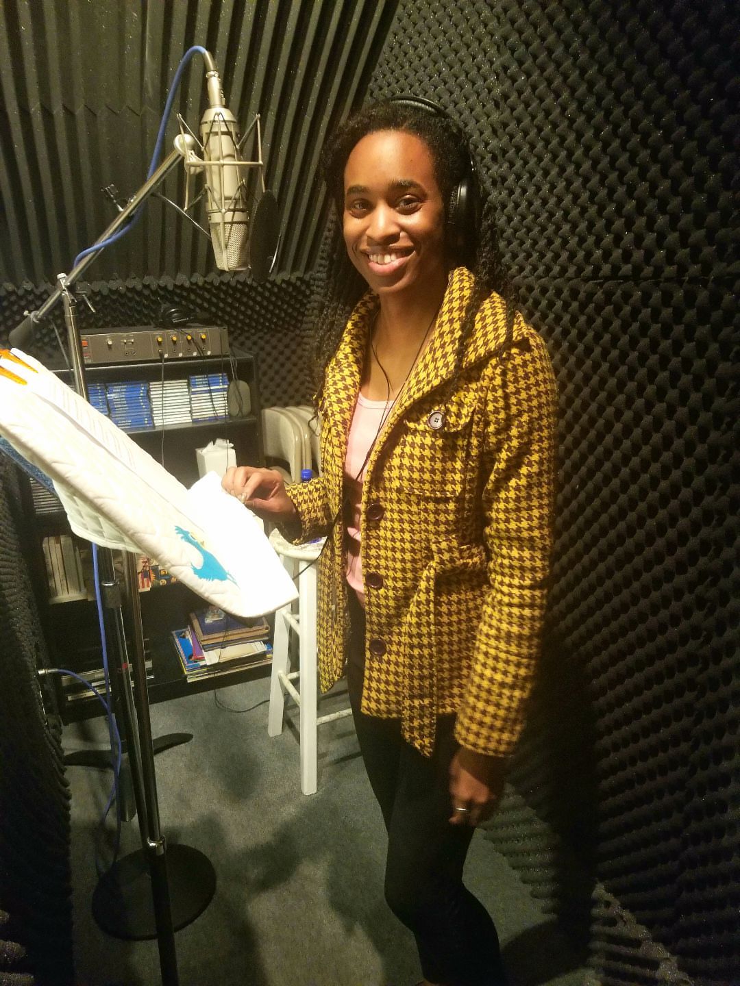 Woman in recording studio with headphones, standing near microphone and music stand, smiling, black soundproof walls behind her.