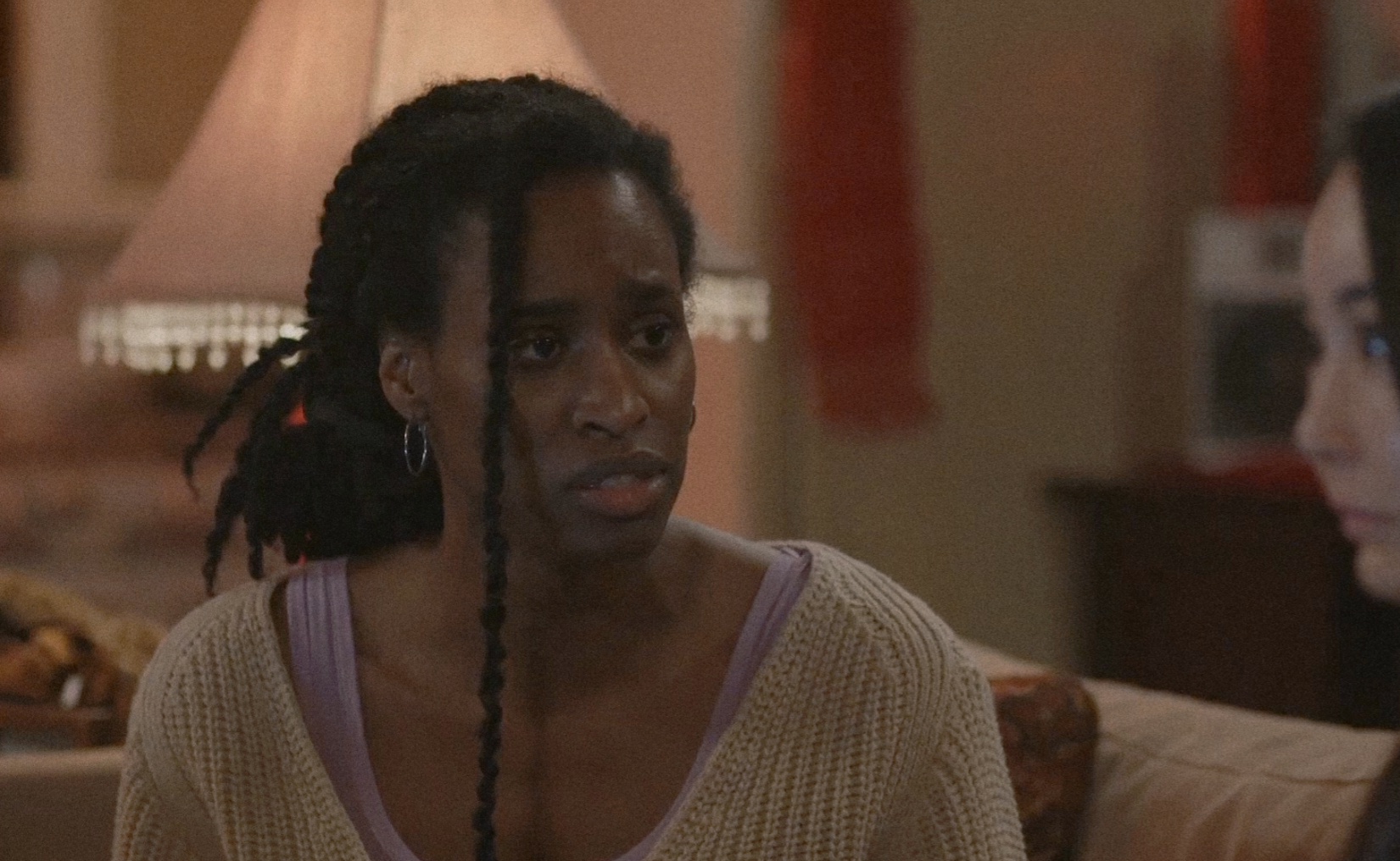 Woman with braided hair looks concerned, sitting indoors with a lamp and furniture in the background.