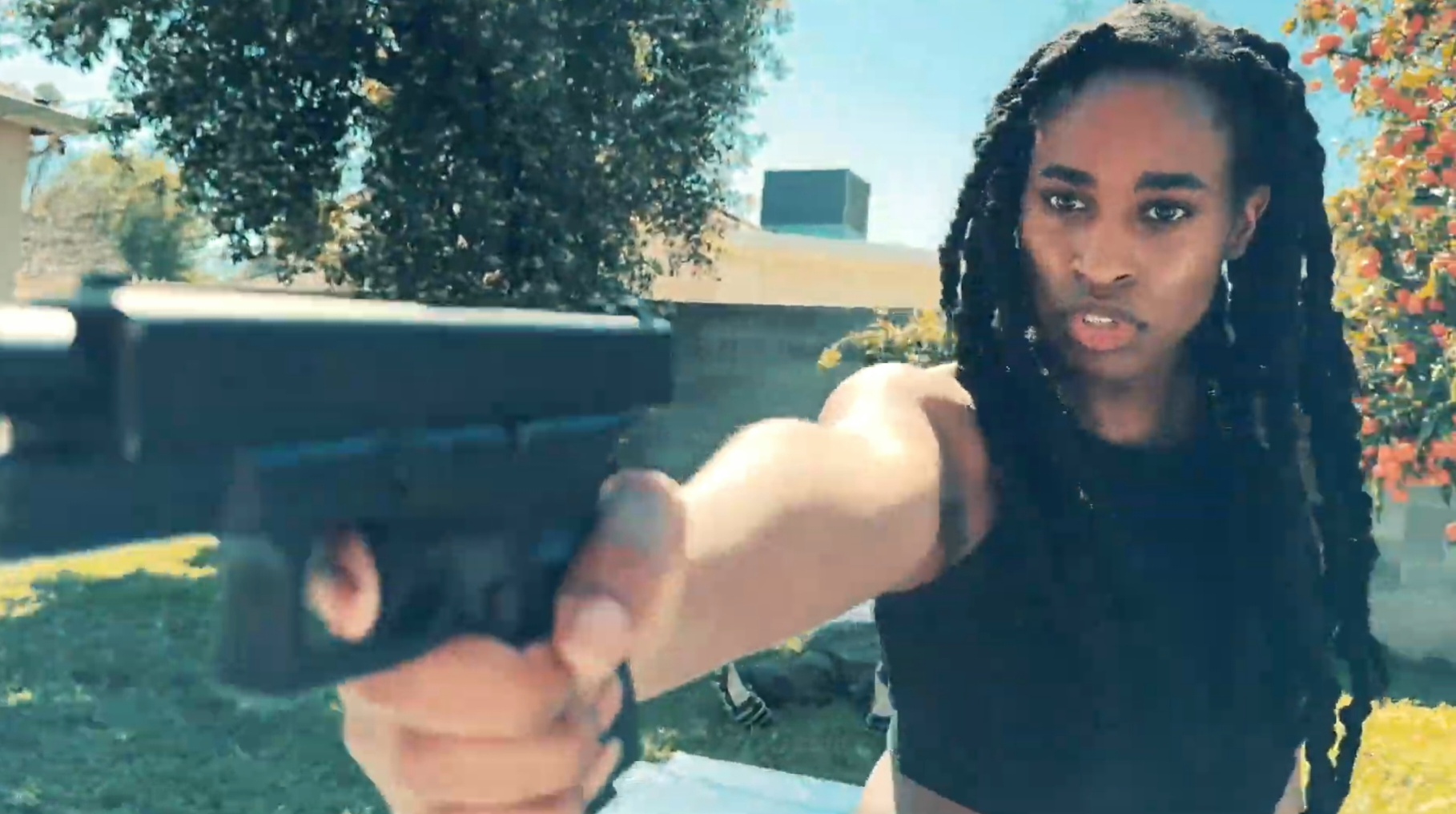 Woman with dreadlocks pointing a gun outdoors, trees and buildings in background.