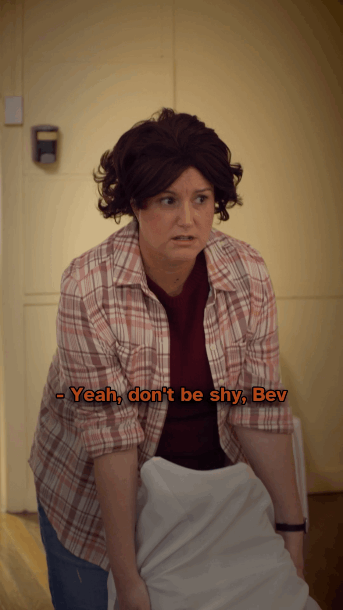 Woman with curly hair wearing a plaid shirt leaning forward, with a serious expression, in a room with yellow walls.