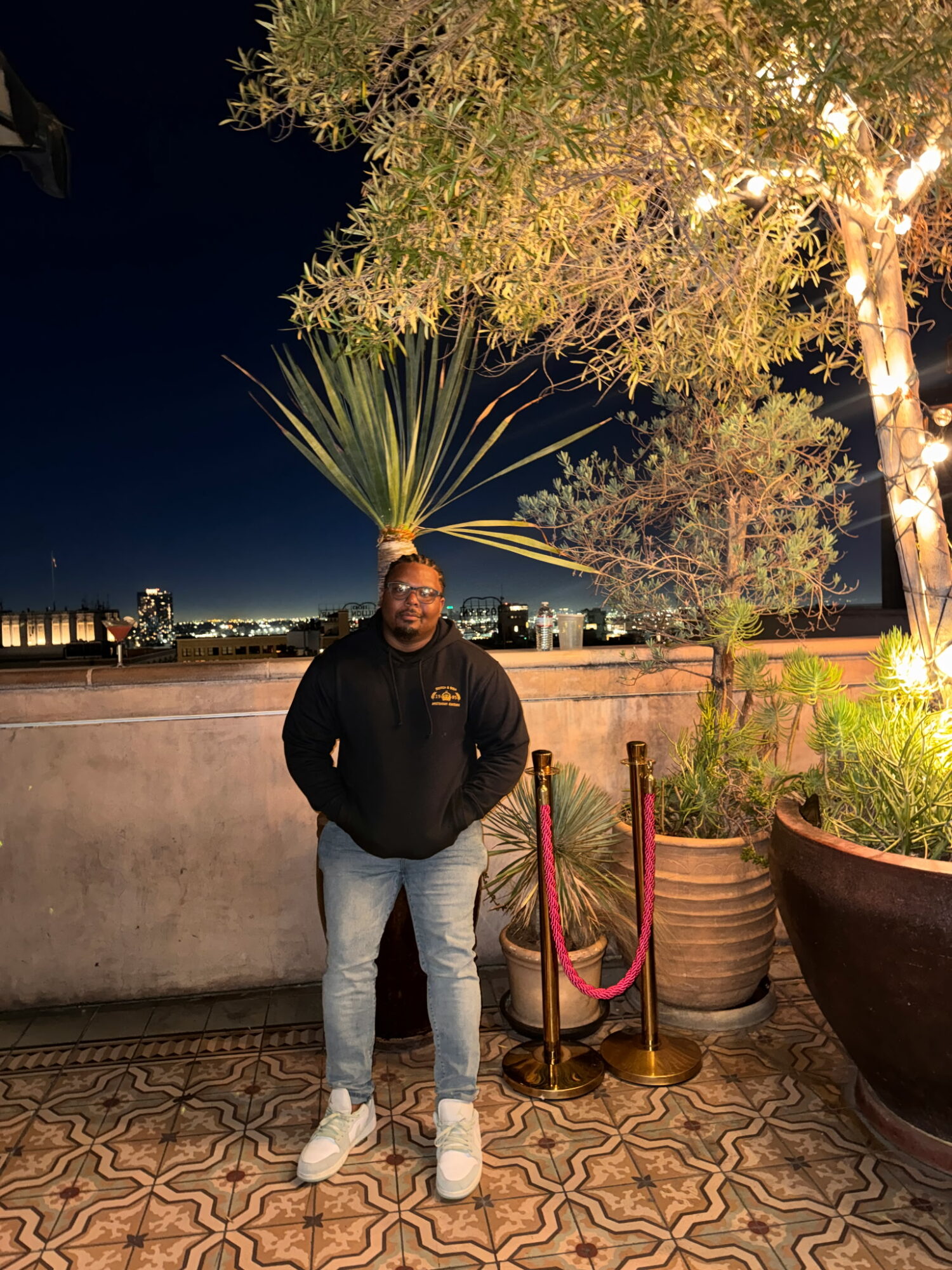 Person standing outdoors at night near potted plants and a tree, illuminated by warm lighting, with city skyline in background.