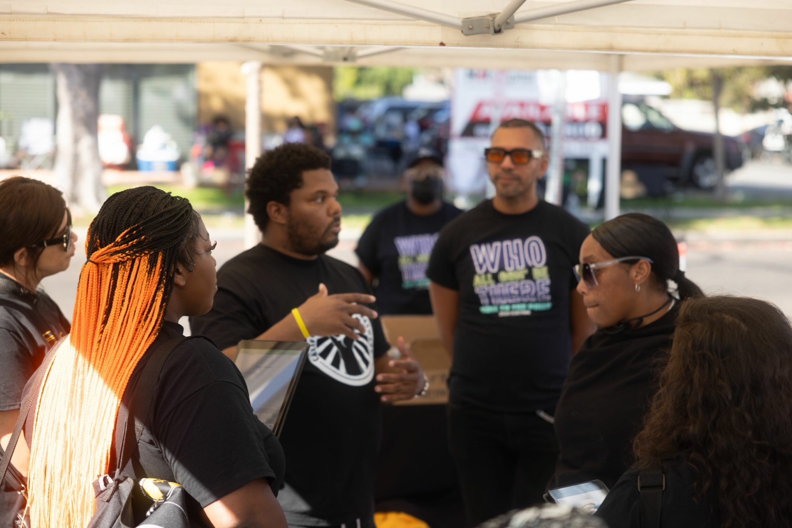Group of people gathered outdoors under a tent, engaged in conversation, with trees and cars in the background.