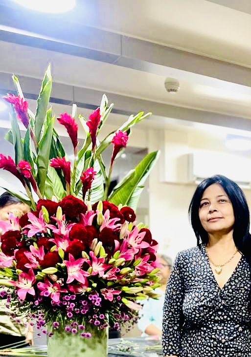 Woman with black hair and patterned dress standing near a large colorful flower arrangement indoors.