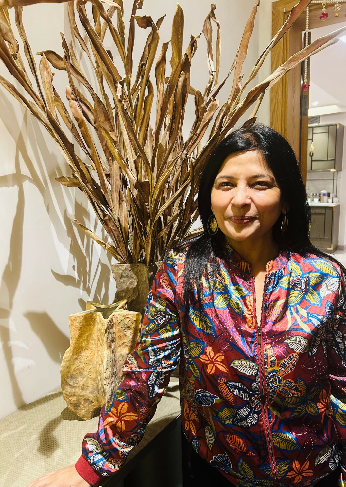 Woman with dark hair wearing a colorful patterned jacket standing next to a large dried plant in a pot, indoors.