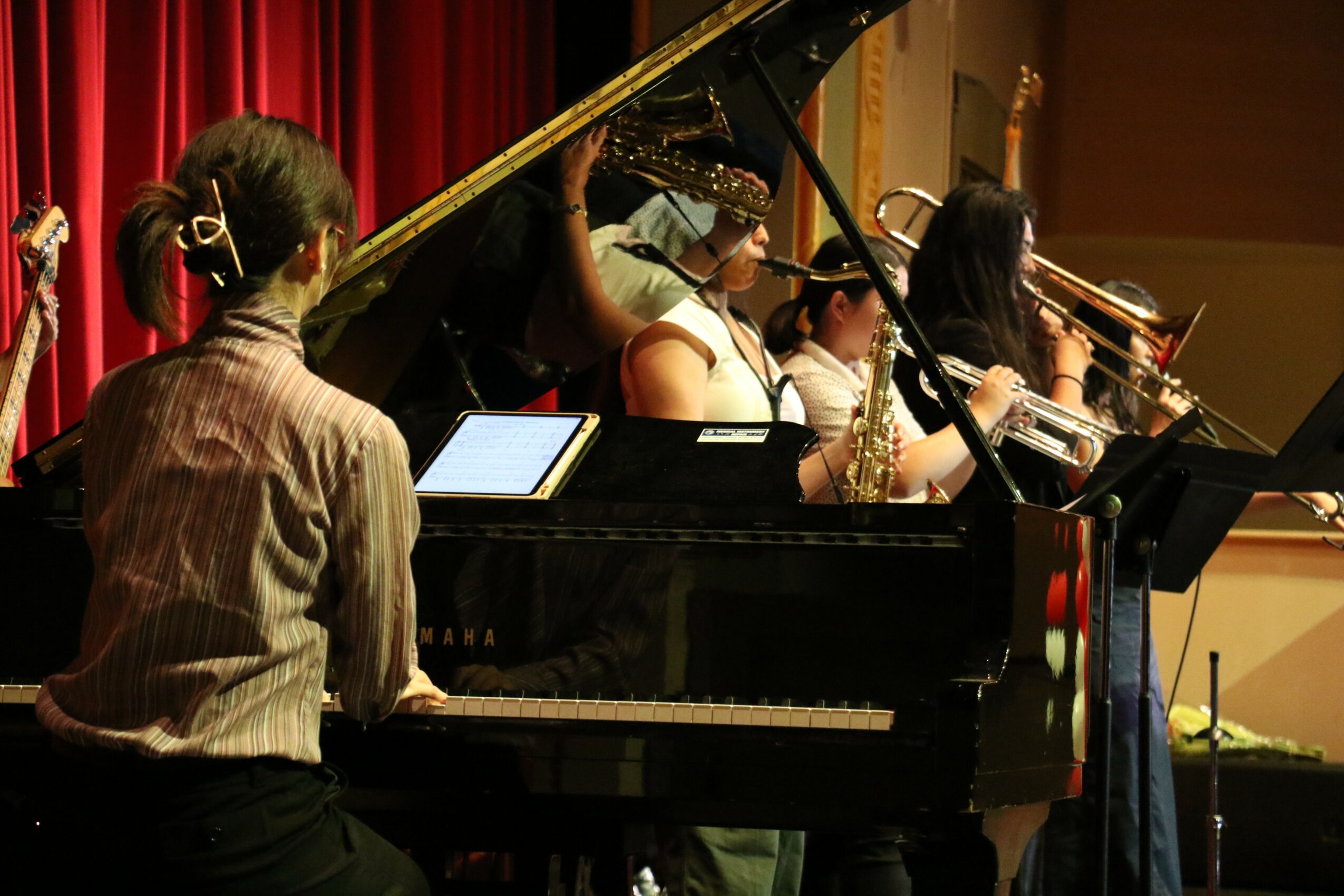 Group of musicians performing on stage with a grand piano and red curtain backdrop, some playing wind instruments.