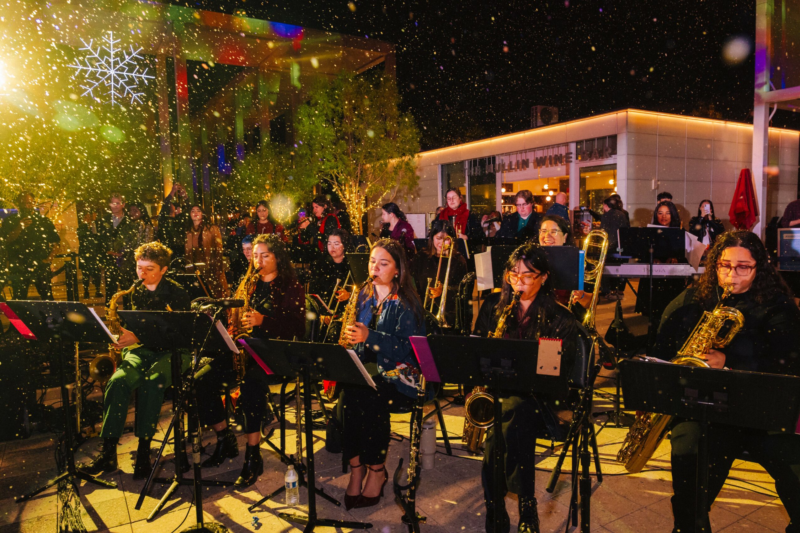 Group of musicians performing outdoors at night with festive lights and snowflake decorations, some wearing masks.