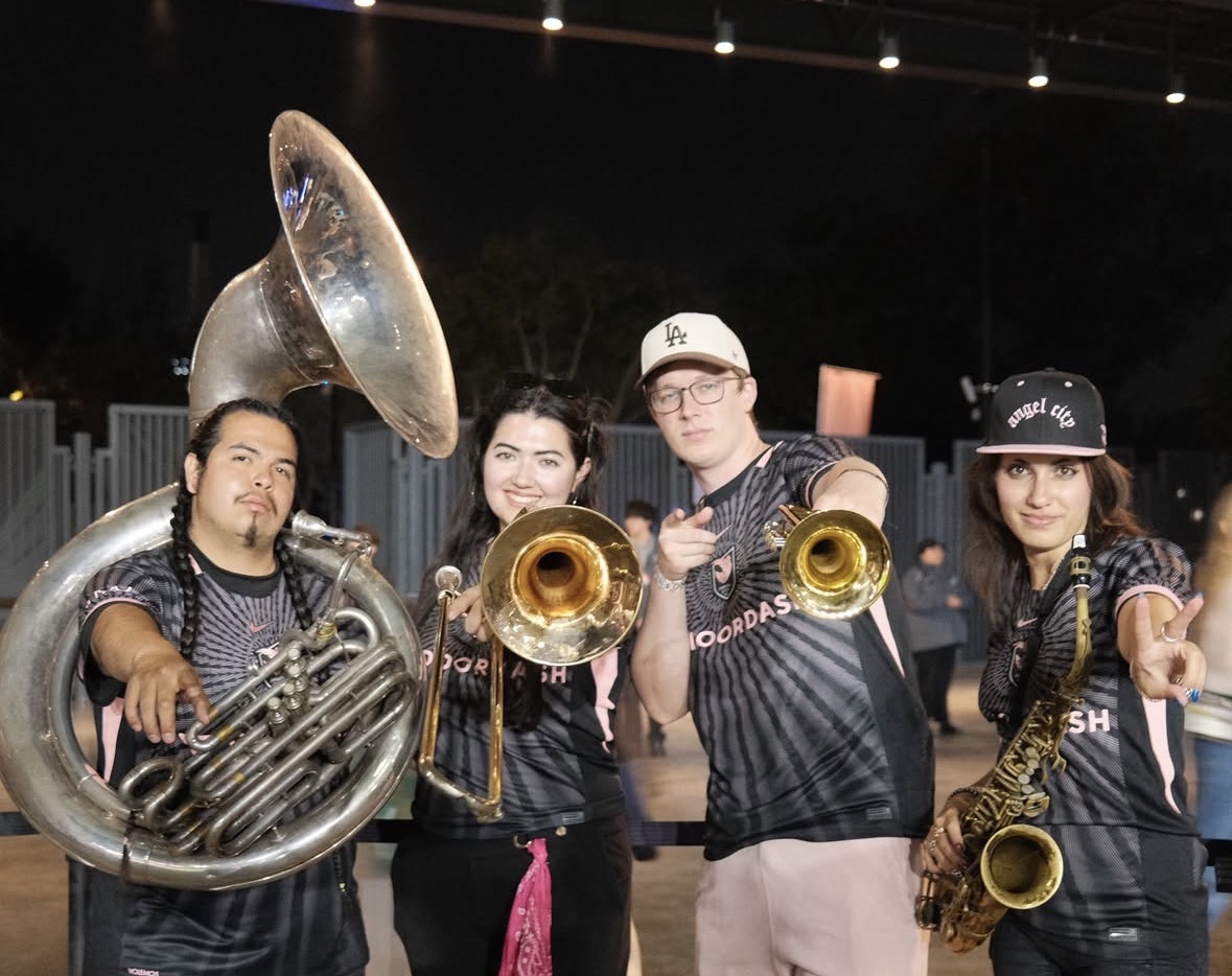 Four musicians holding brass instruments, standing outdoors at night, smiling and pointing at the camera.