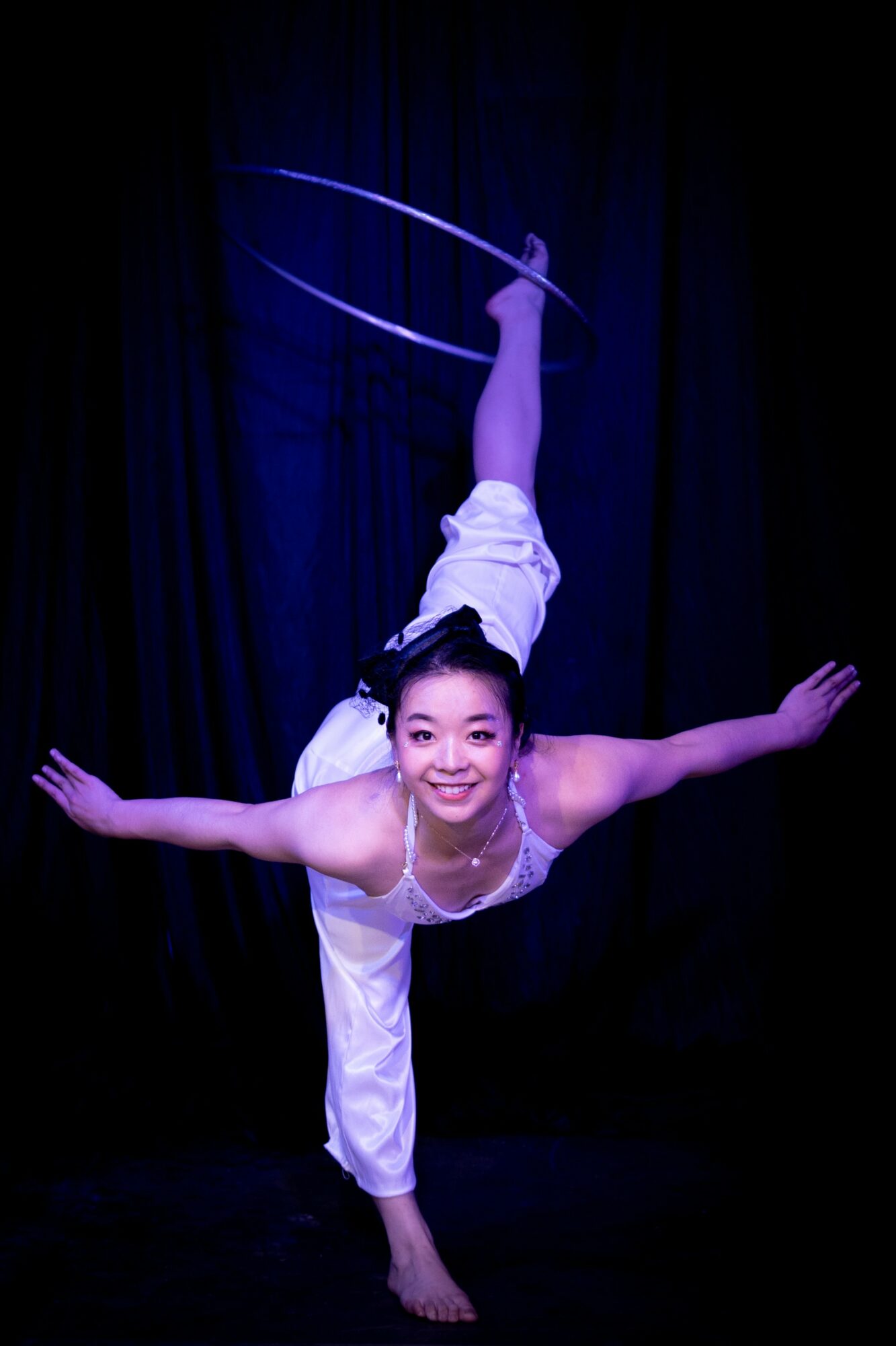 Performer balances on one hand with legs extended upward, holding a hoop, against a dark background.