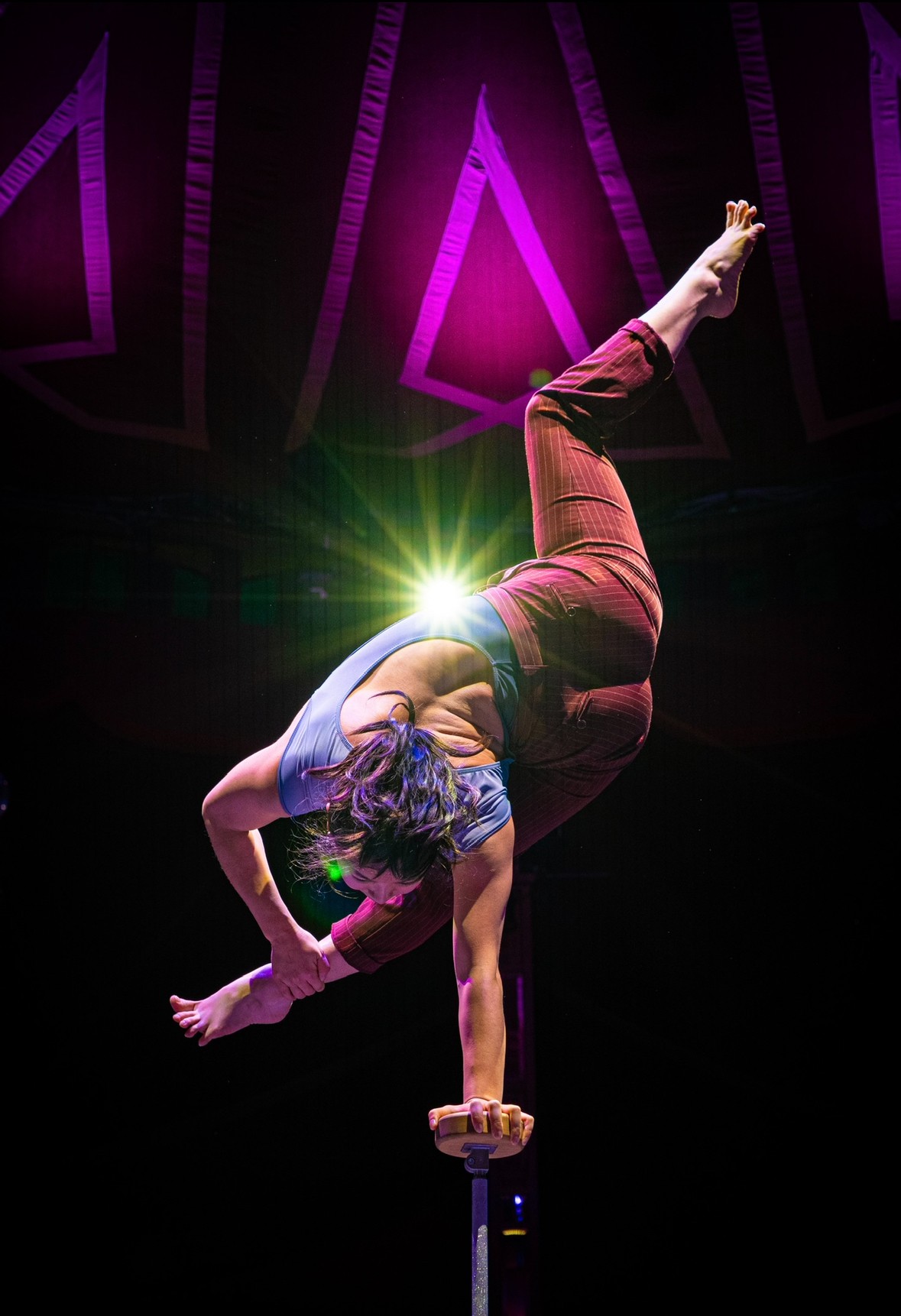 A person performs a handstand on a balance object, with a bright light behind them and purple stage lighting above.