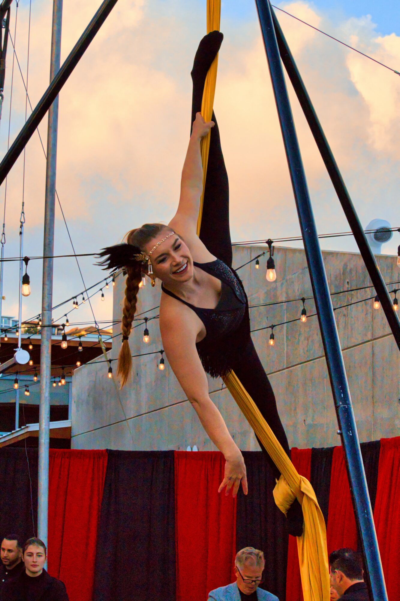 A woman performs aerial silk acrobatics outdoors, hanging upside down with a smile, against a sky with clouds and string lights.