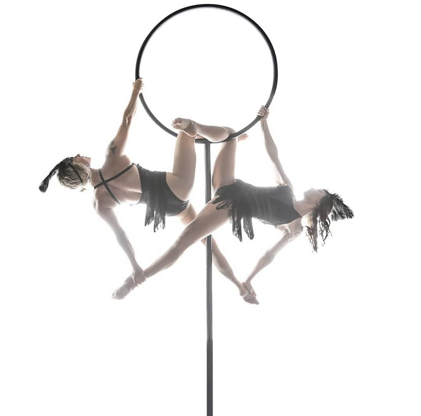 Two performers in black costumes hold a hoop, balancing on a pole with their bodies extended outward.