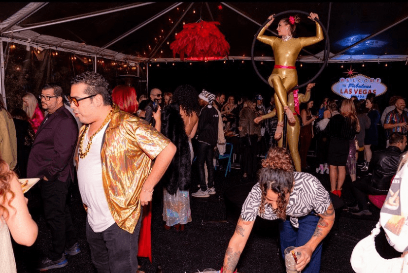 Crowd of people at an event with a large golden statue of a woman holding a hoop, decorated with red feathers, in the background.