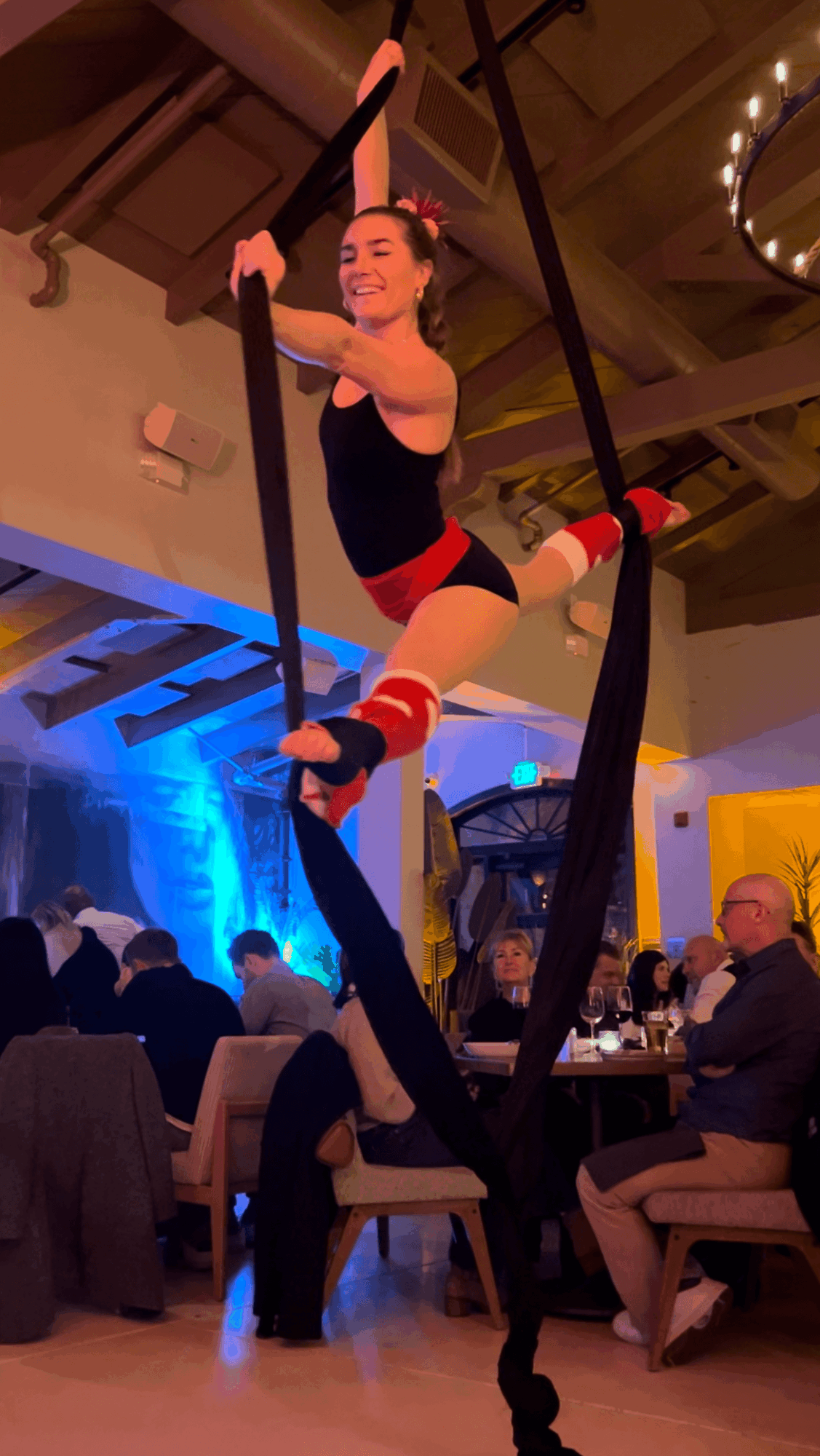 Aerialist performs on black silk fabric in a decorated indoor venue with audience seated at tables.