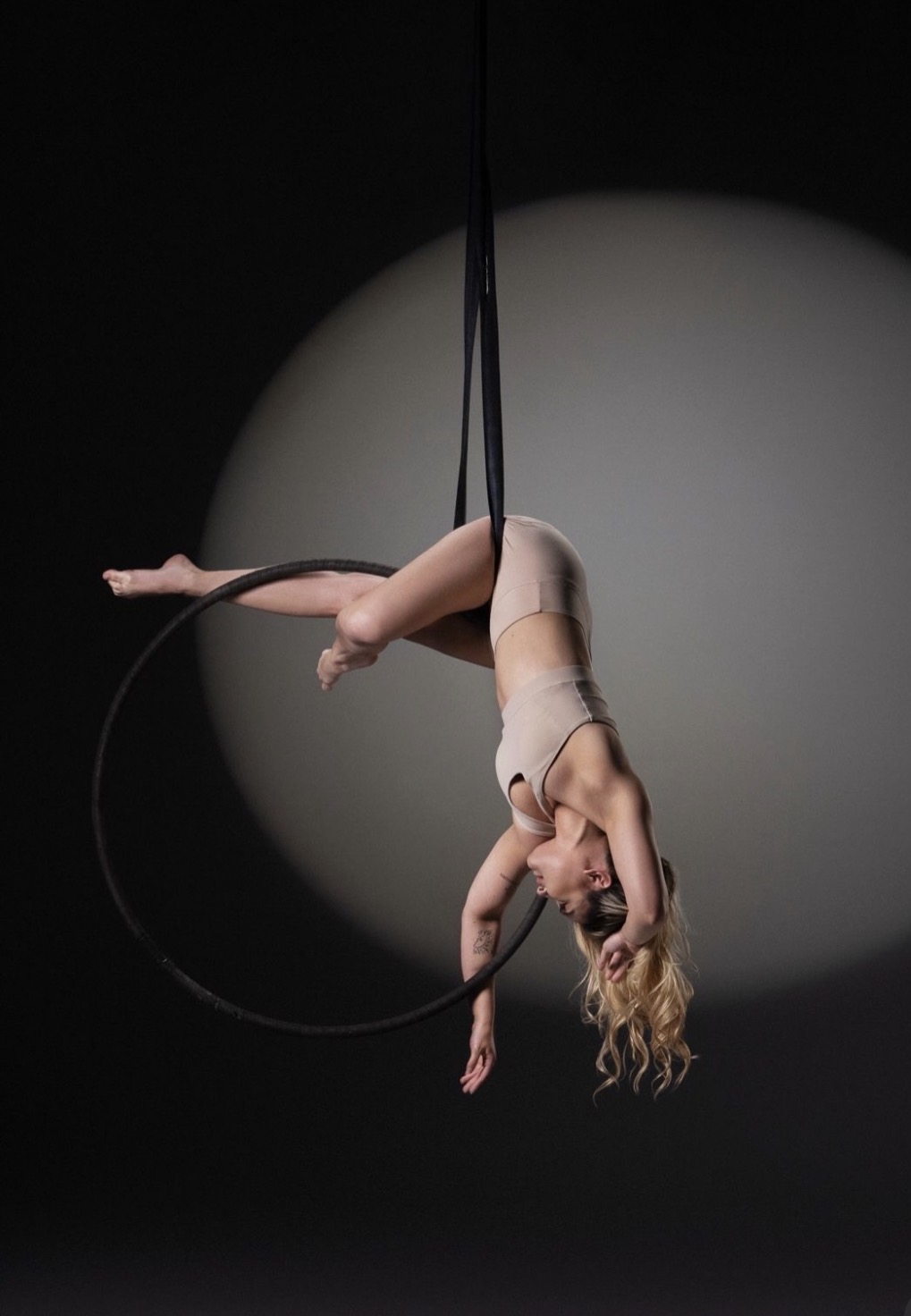 Aerialist performs on a hoop, hanging upside down with her hair hanging downward, against a dark background with a spotlight.