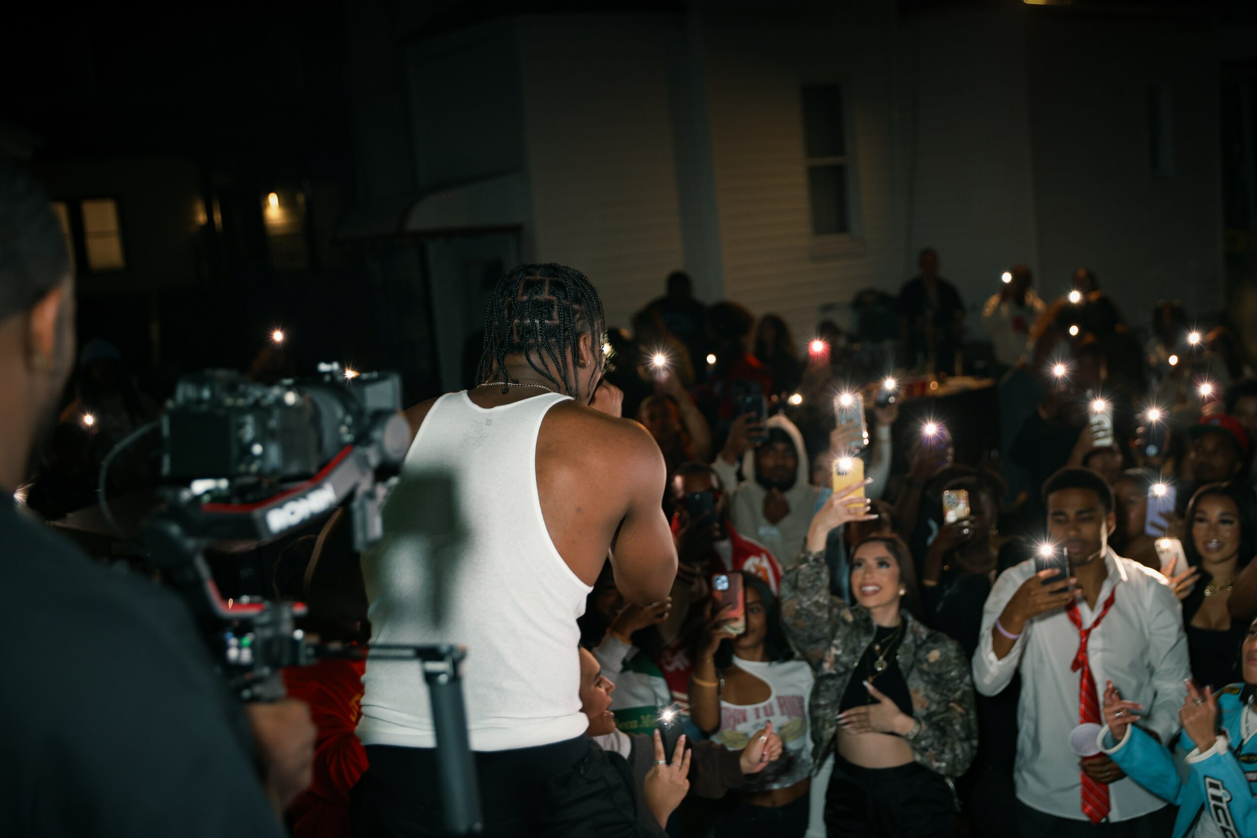 Crowd of people at night with many holding up phones with flashlights on, some smiling and taking photos.