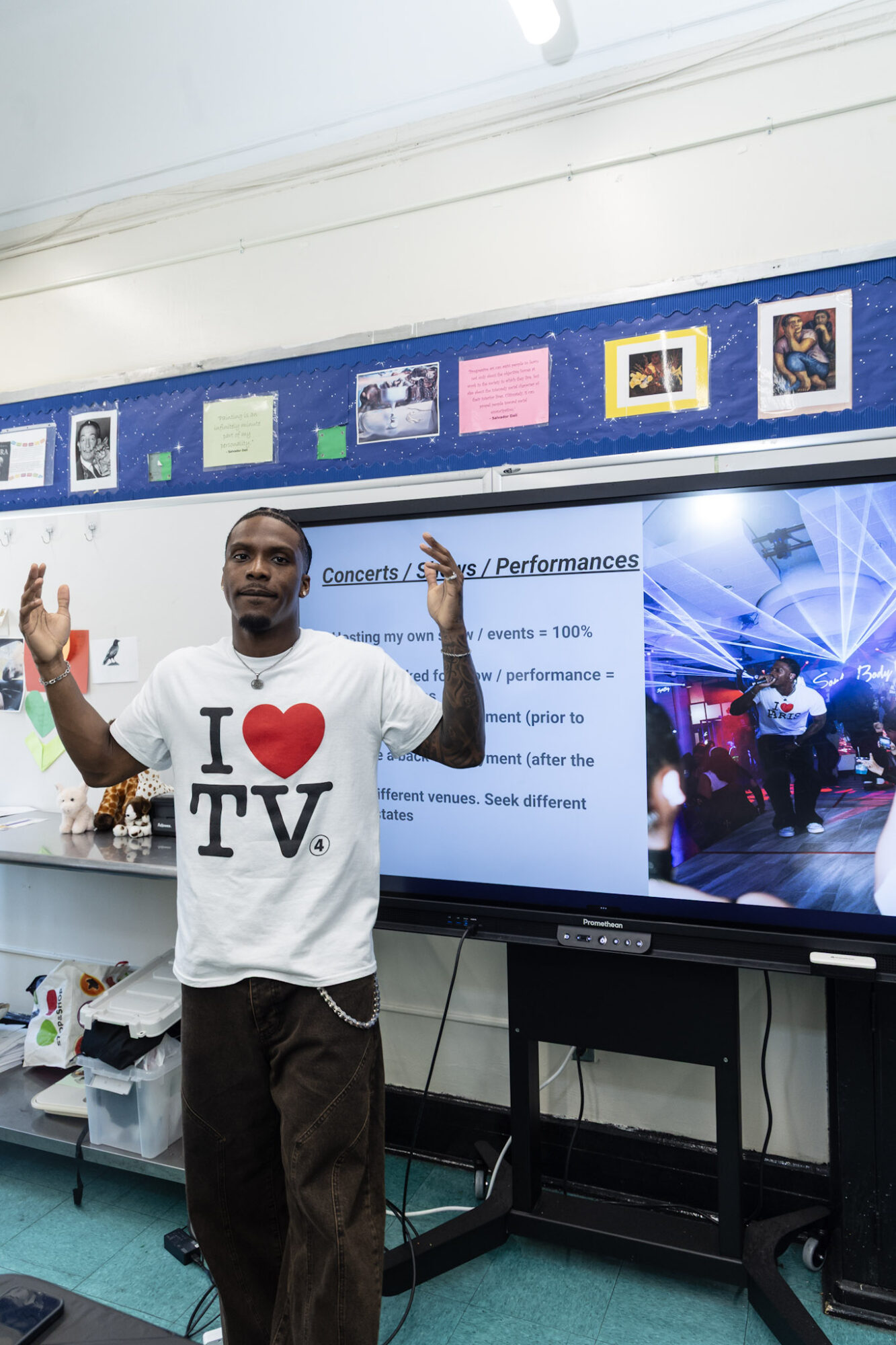 Person standing in front of a large screen in a classroom, wearing a white T-shirt with 'I ❤️ TV' and raising one hand.