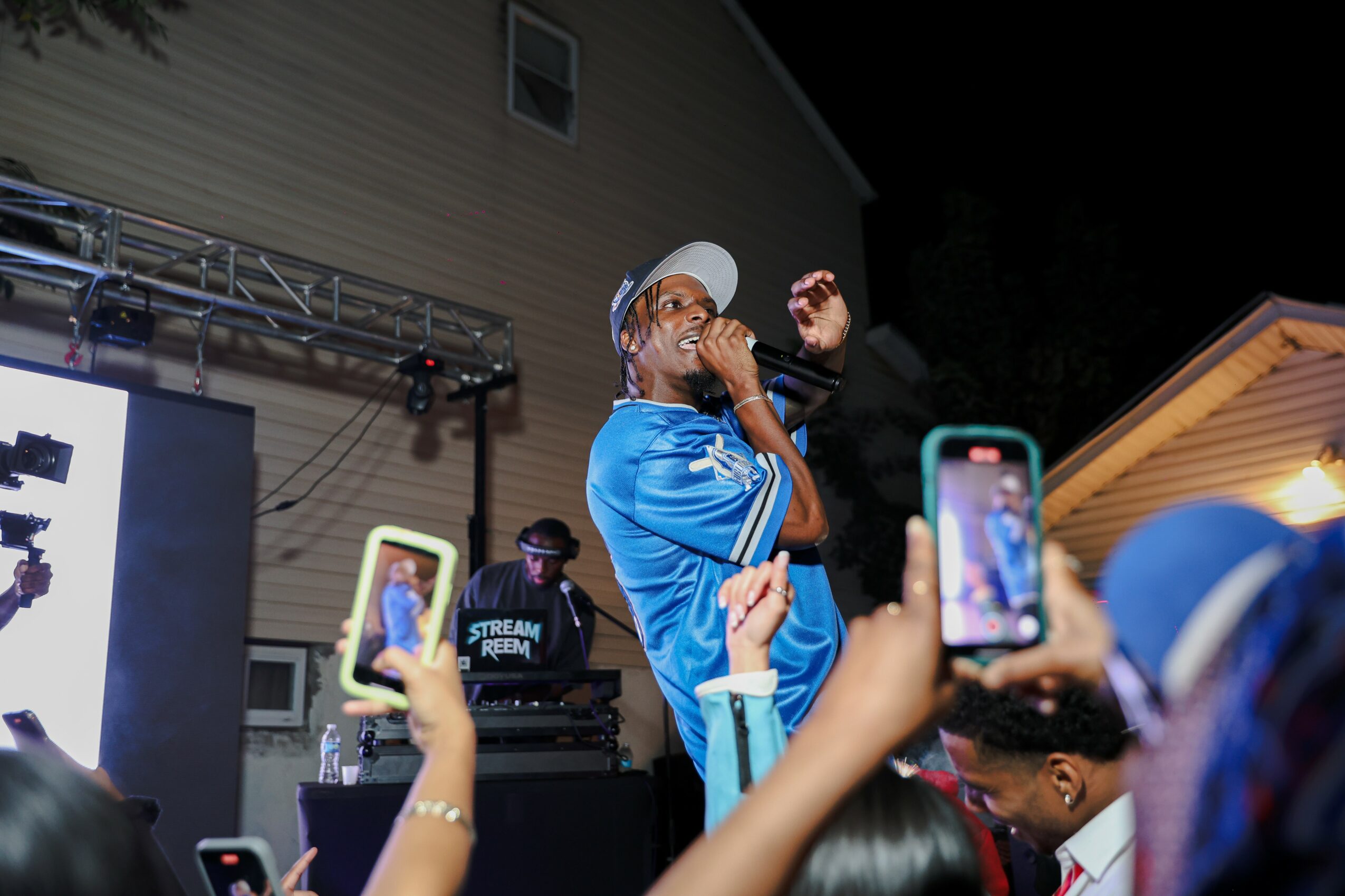 Performer in blue outfit and white hat singing on stage at night, audience taking photos, house in background.