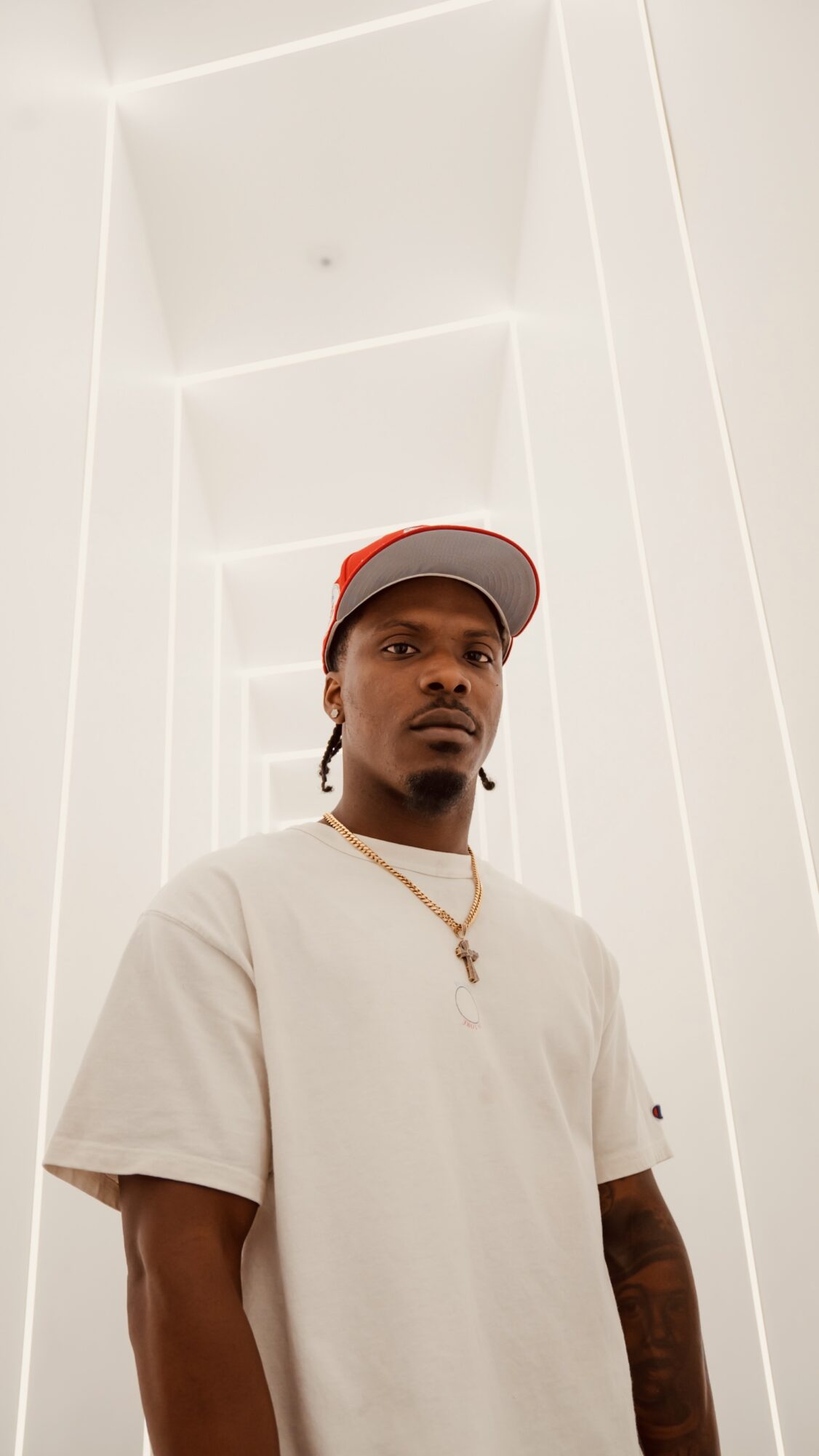 Young man wearing a white t-shirt, red and white cap, and a gold chain with a cross, standing indoors.