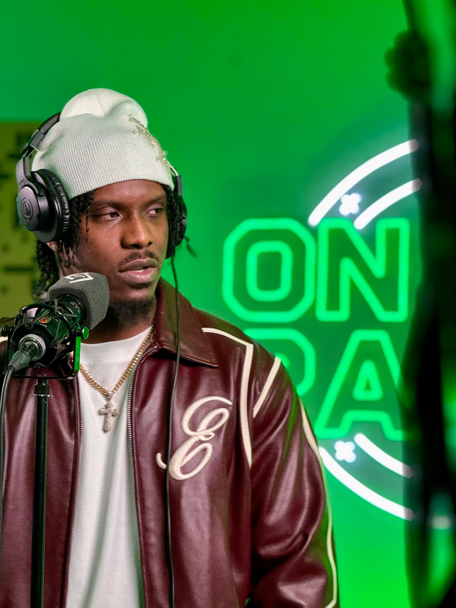 Person with headphones and white beanie in front of a green background with a neon sign reading ON AIR.