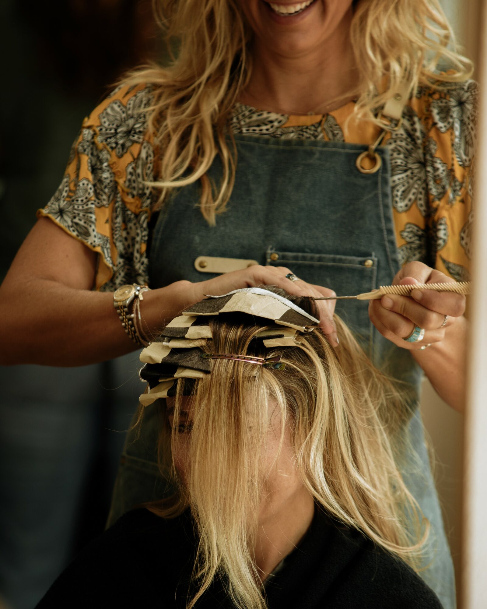 Person with blonde hair getting hair styled with rollers, wearing a patterned shirt and apron, smiling.