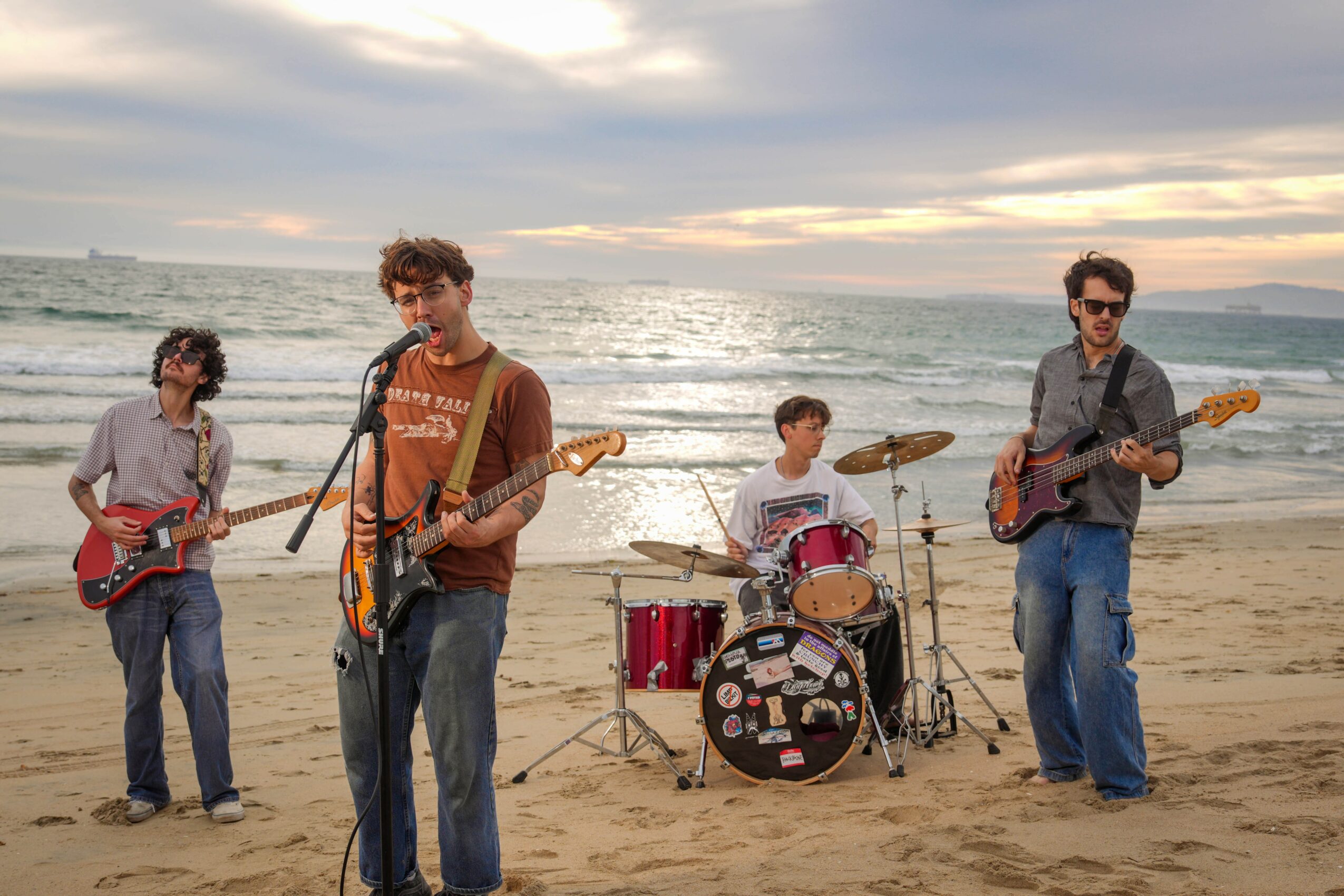 Four musicians perform on a beach at sunset, playing guitars and drums, with ocean waves in the background.