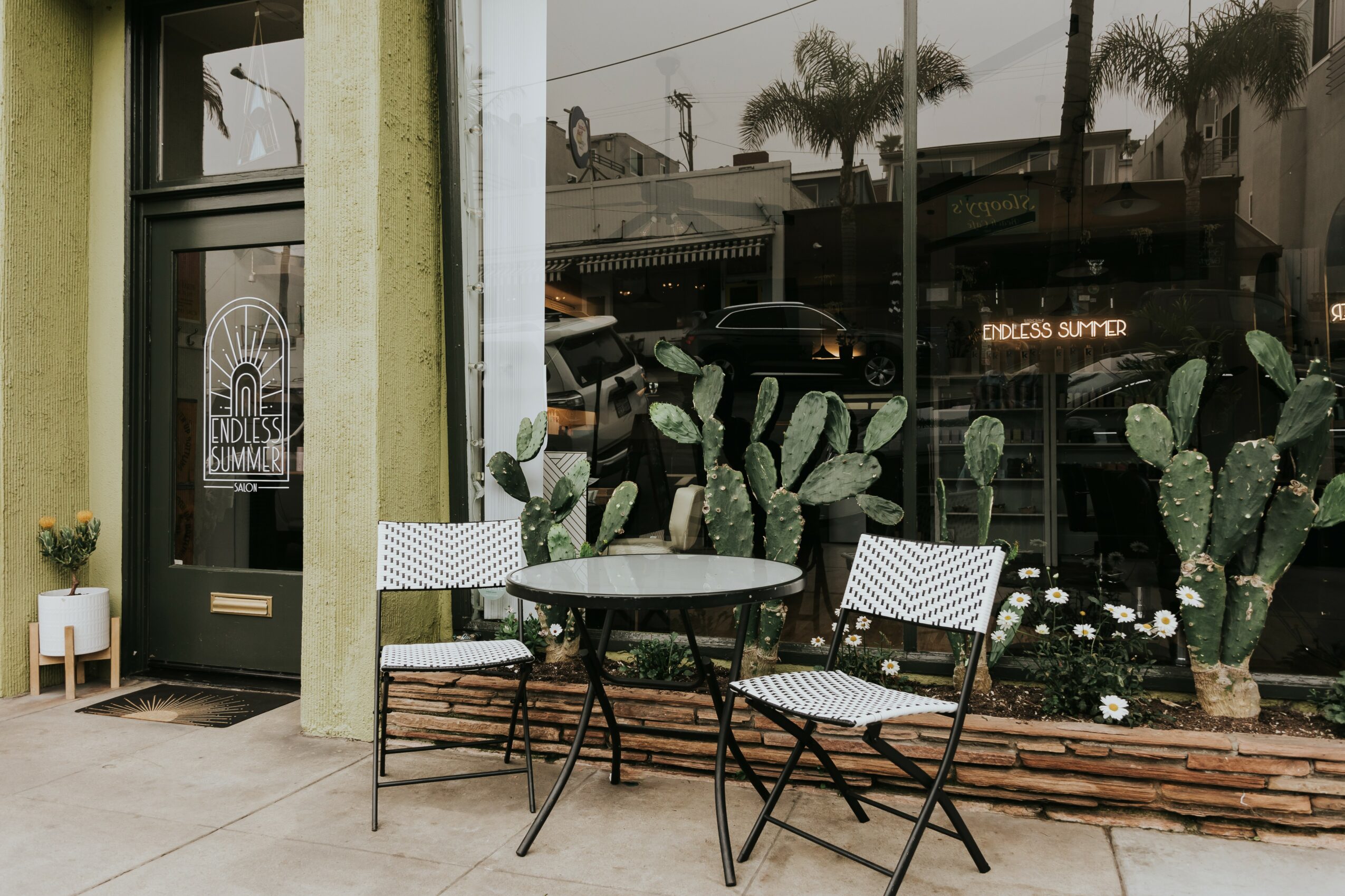 Outdoor seating with a round table and two chairs in front of a large glass window with cactus plants inside, sidewalk nearby.