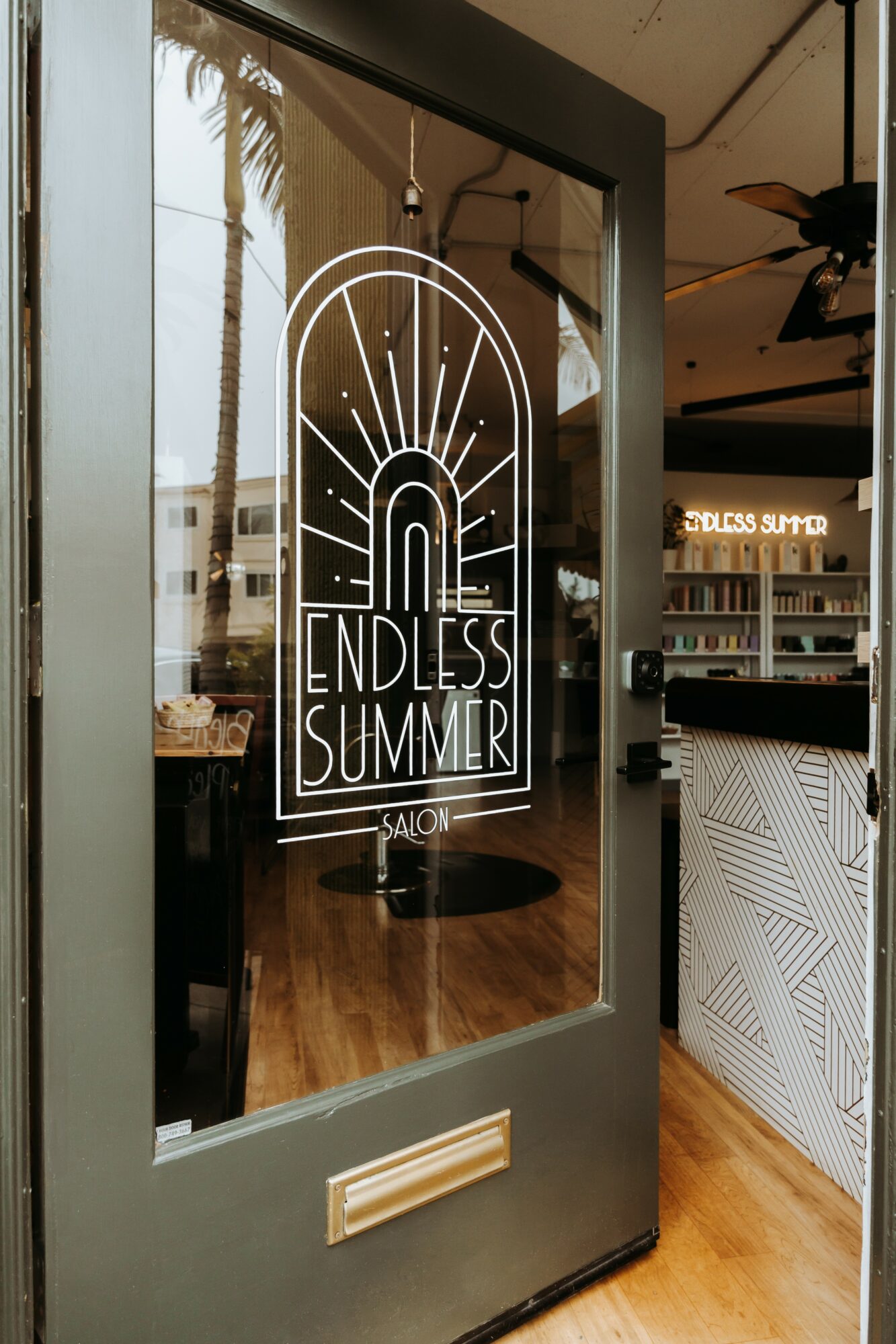 Glass door with a neon sign reading 'Endless Summer' and a sunset graphic, inside a modern cafe or salon.