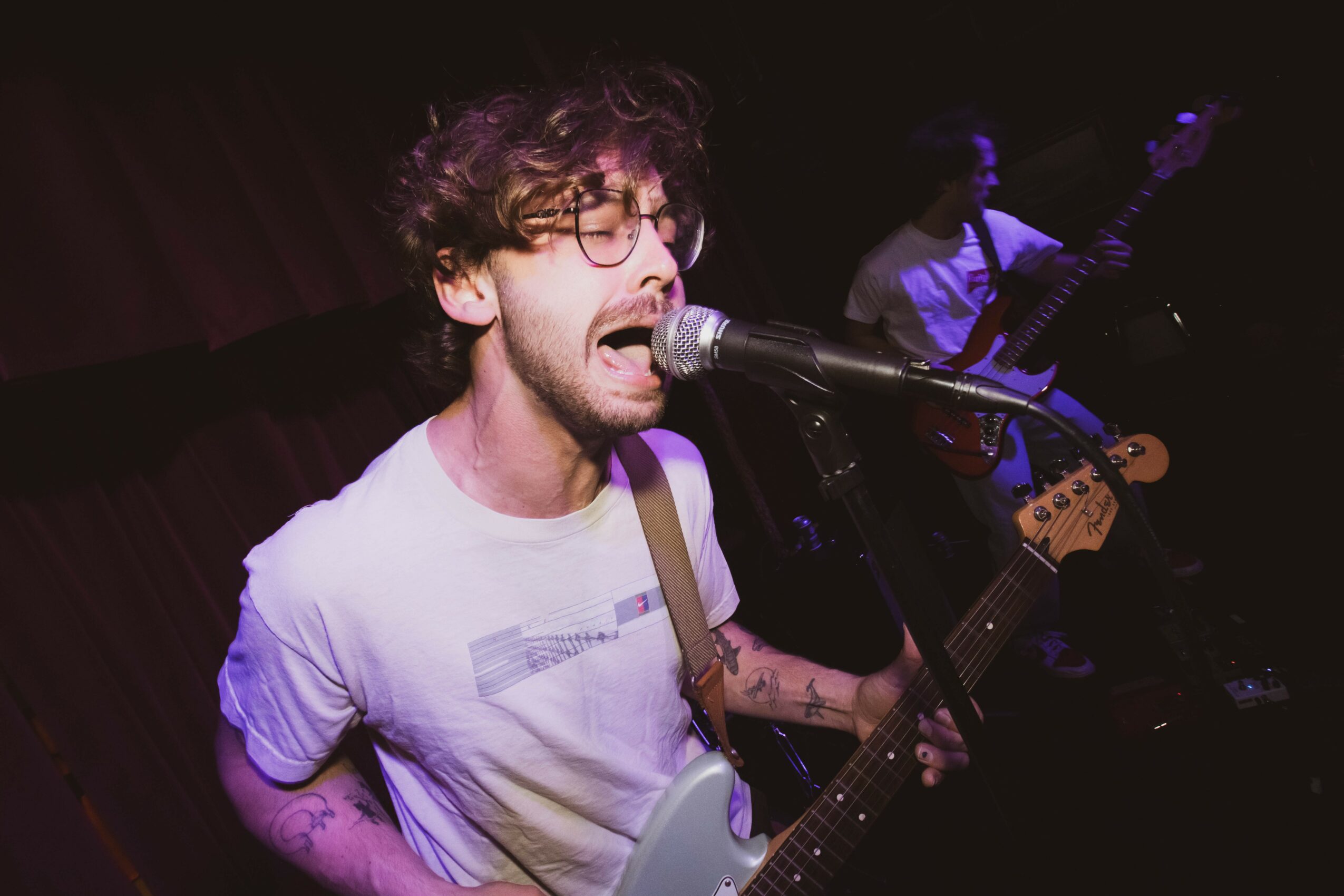 A musician with glasses and curly hair sings into a microphone while playing an electric guitar on stage.