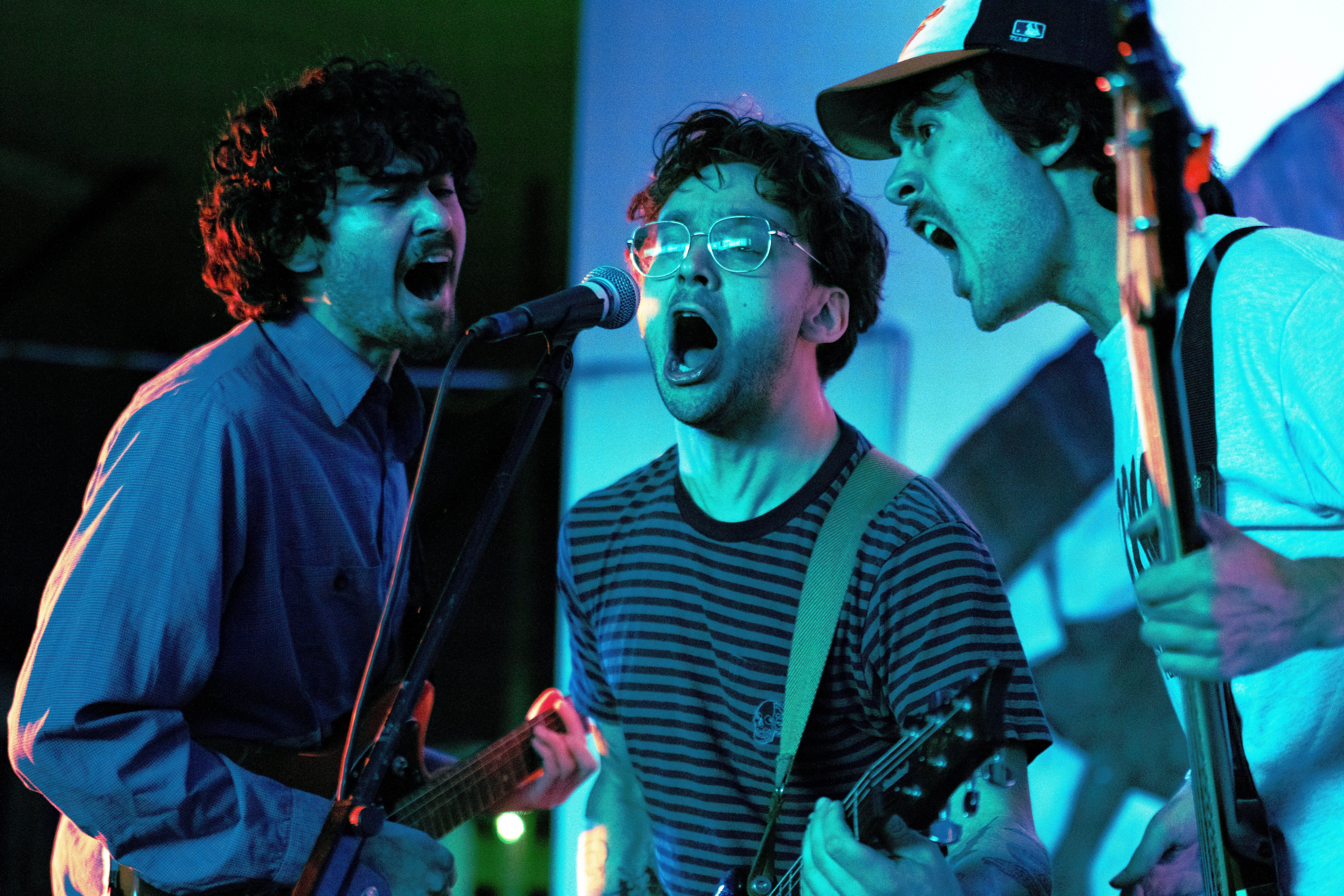 Three people singing passionately into a microphone, playing guitars, with colorful stage lighting in the background.