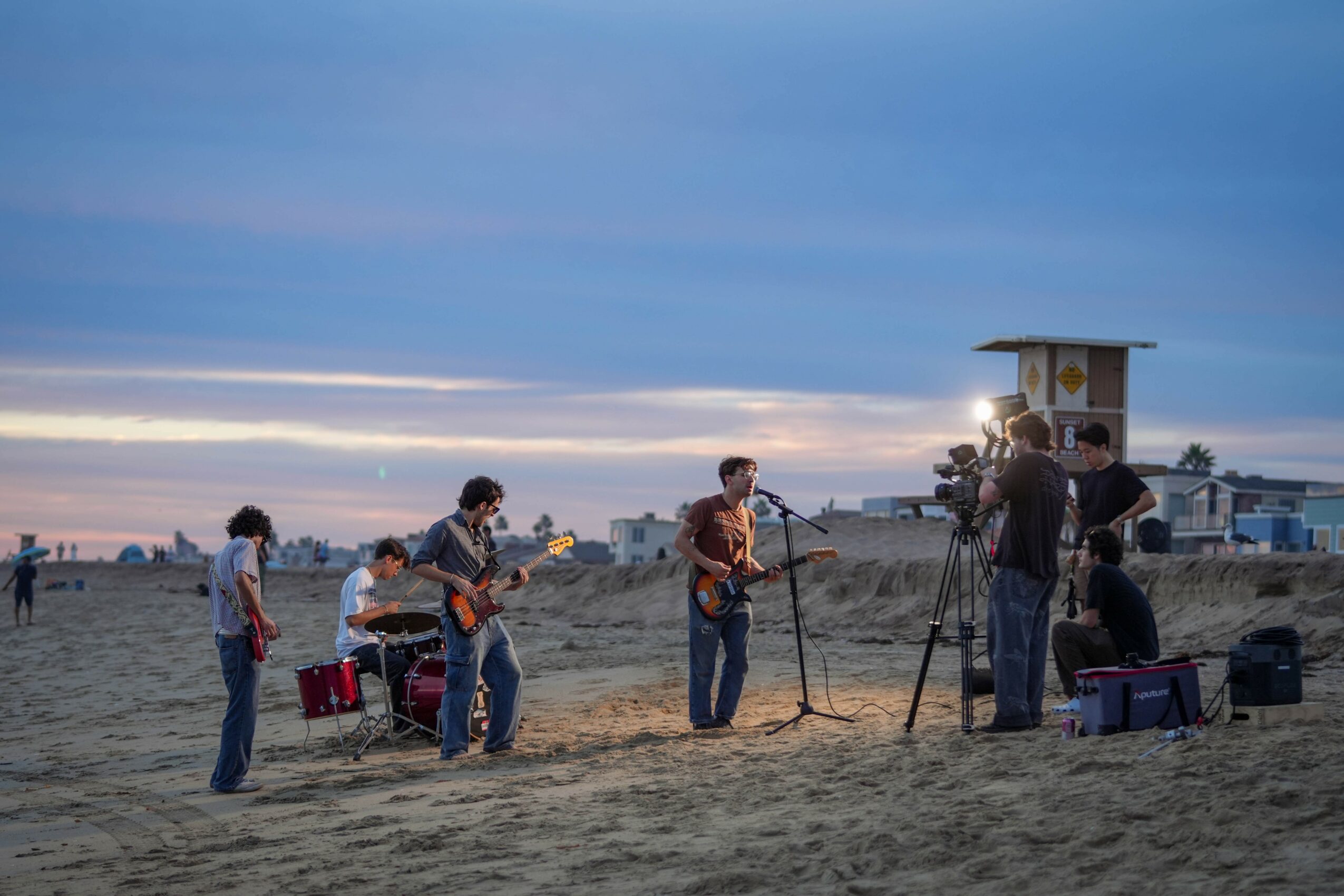 A group of musicians performing on a beach at sunset, with people watching and equipment nearby.