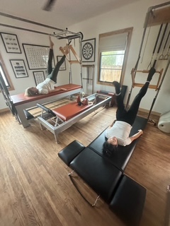 Two women exercising on a wall-mounted bar and a mat in a room with wooden floor and framed pictures on wall.