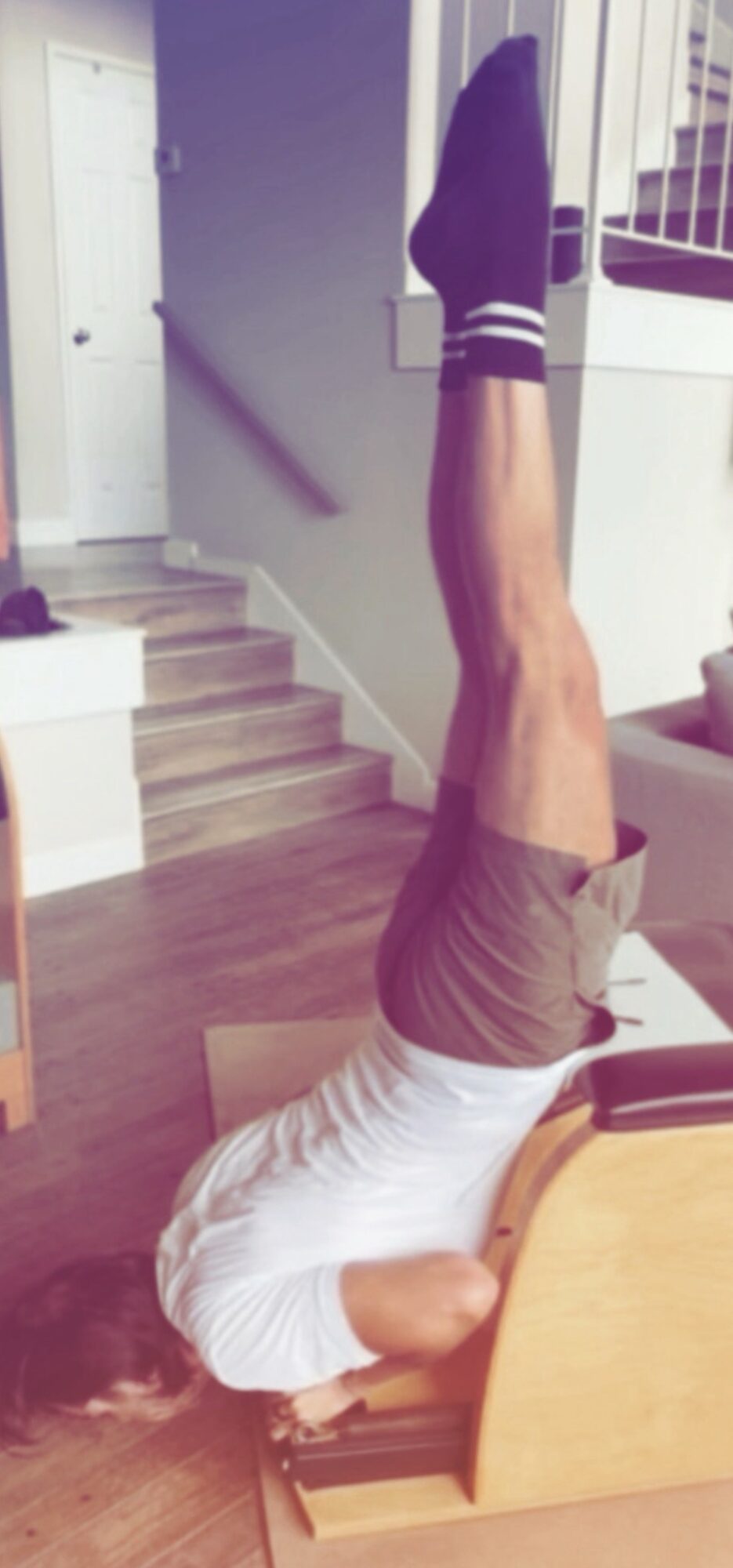 Person lying upside down on a chair with legs extended upward, in a room with stairs and a staircase railing visible
