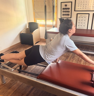 Person lying on stomach on a massage table, reaching forward with arms, in a room with framed pictures and a window.