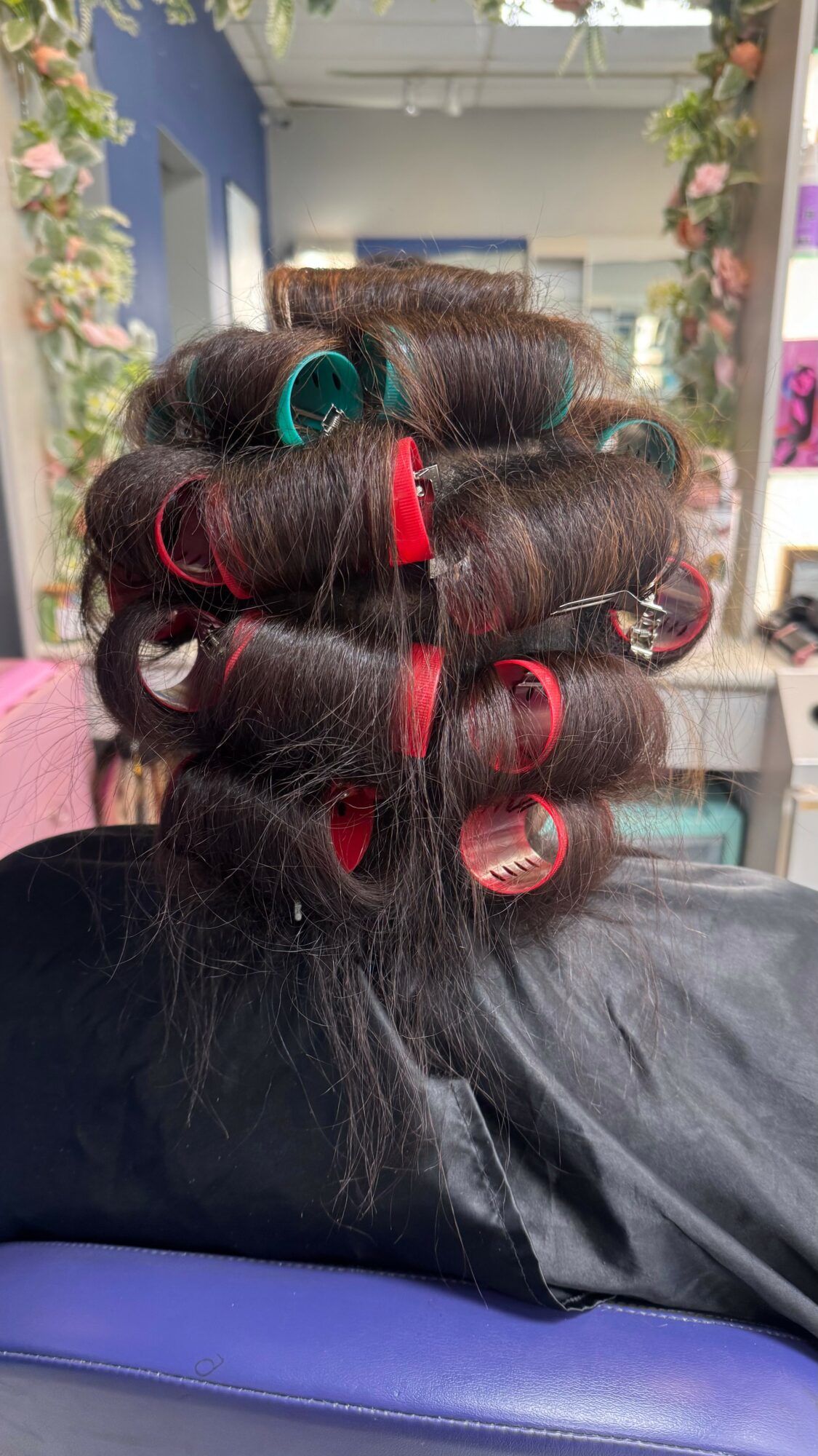 Person with hair rollers in a salon, facing away, with a mirror and salon decor in the background.