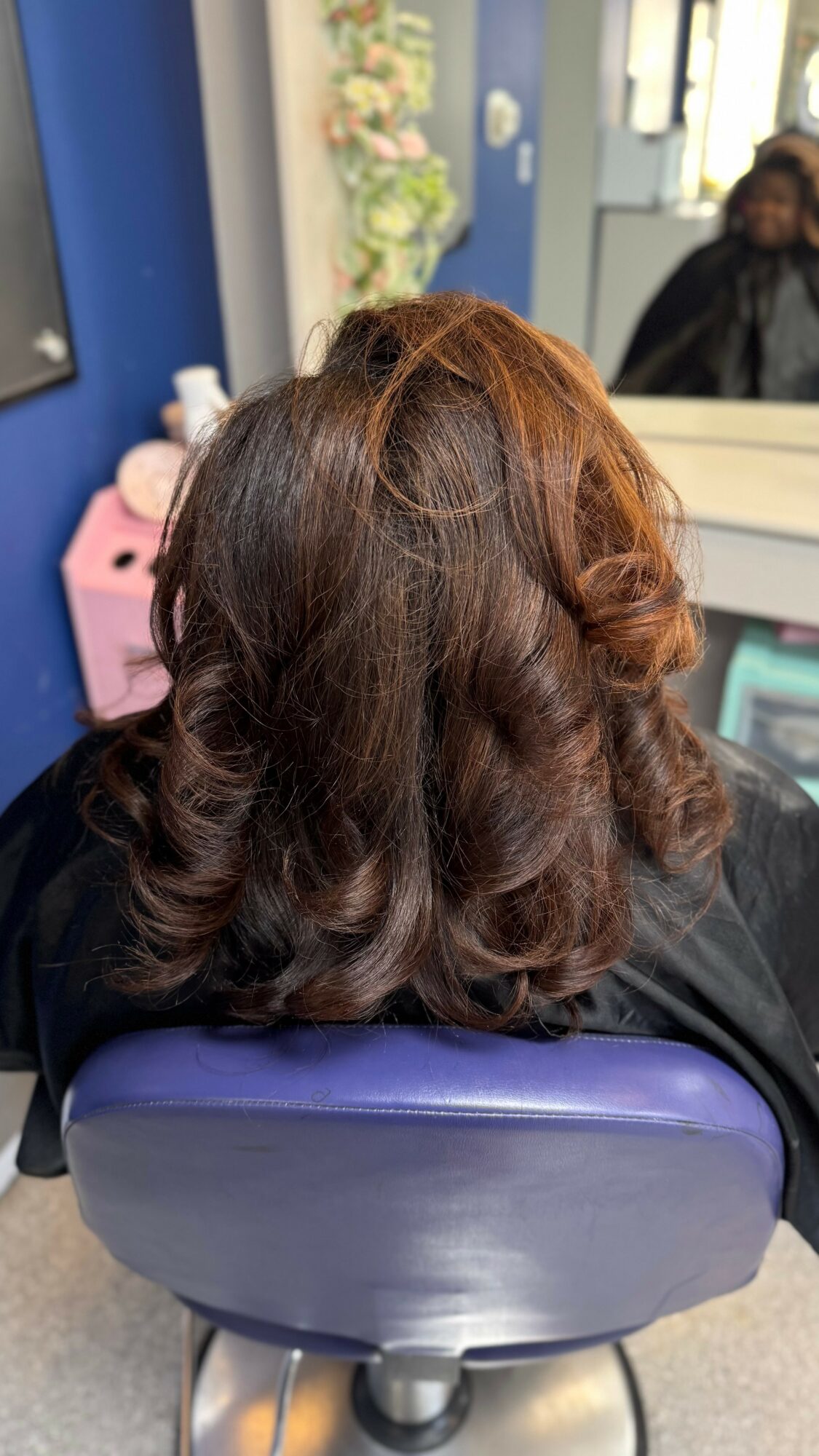 Back view of a woman with shoulder-length, wavy brown hair sitting in a salon chair, mirror and salon tools visible in background.