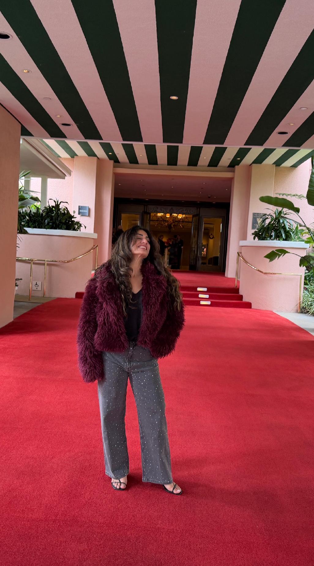 Woman standing on red carpet in front of building entrance with plants on sides, ceiling with black and pink stripes above.