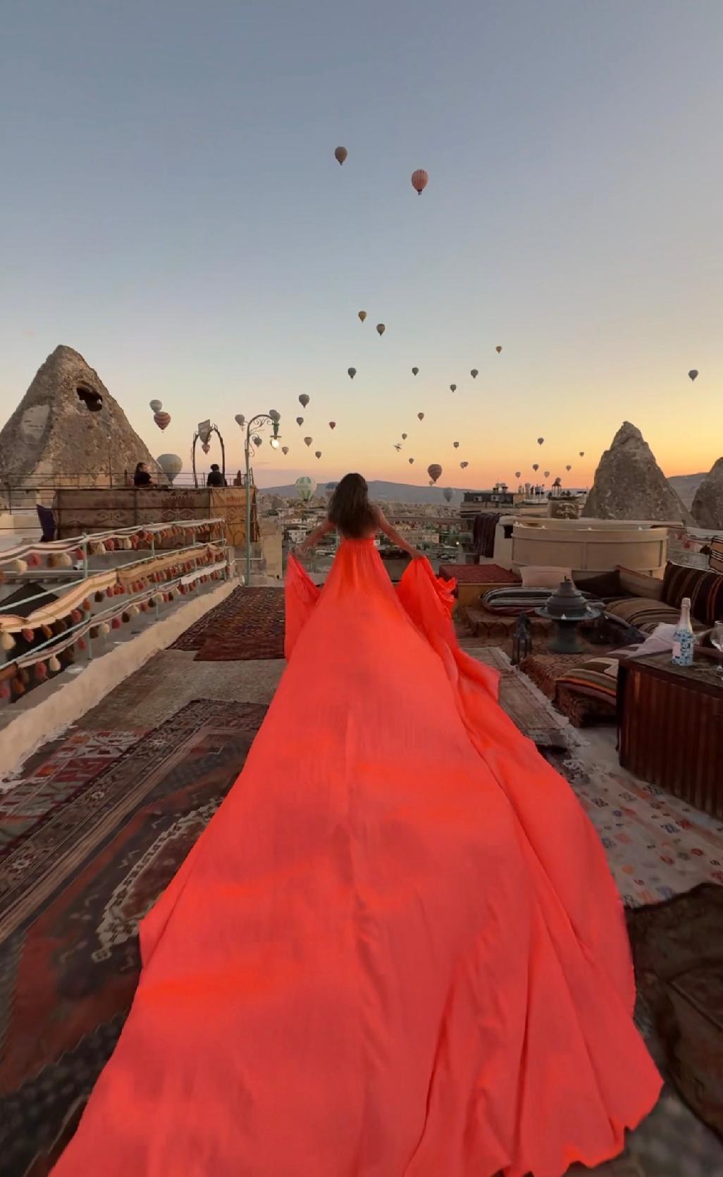 Person in a flowing red dress standing on a rooftop at sunset, hot air balloons in the sky, rocky formations nearby.