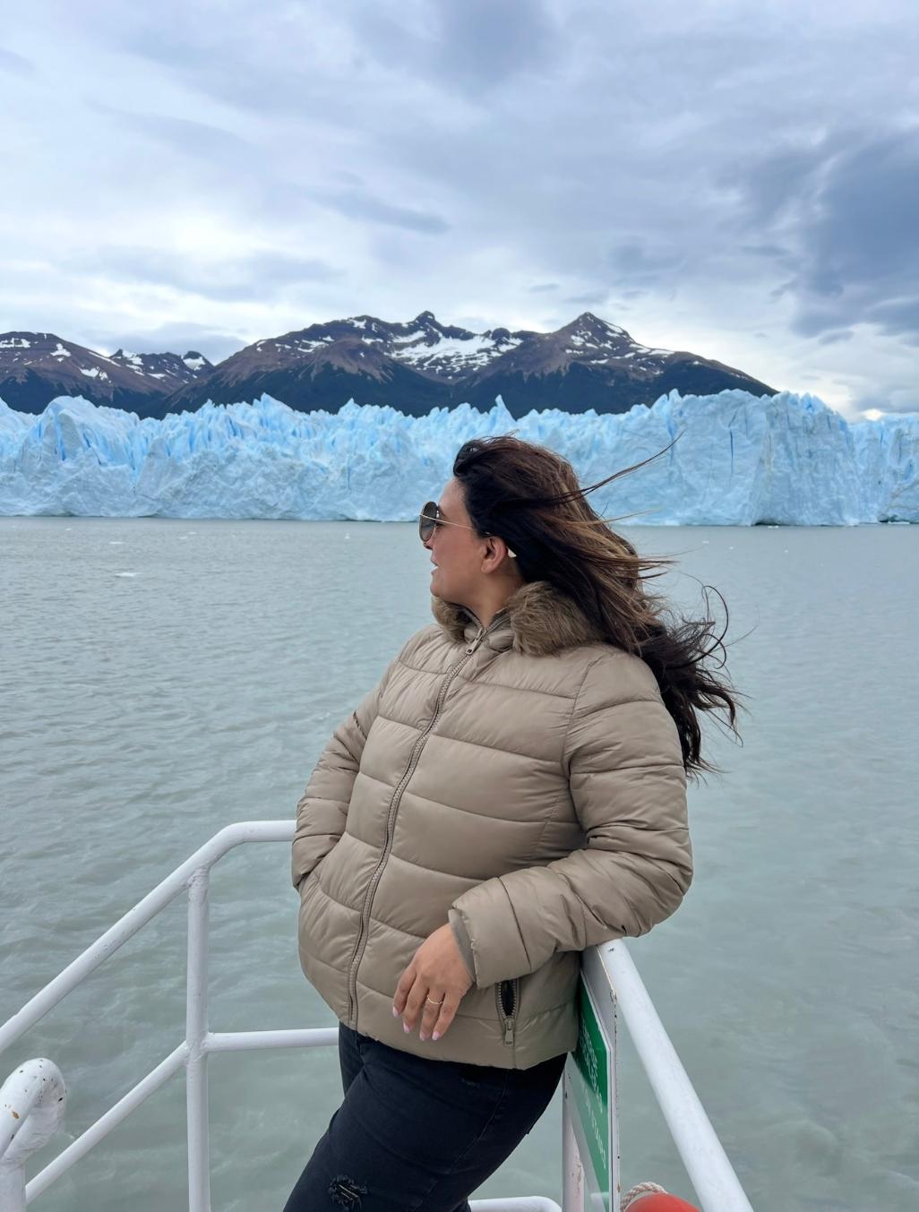 Woman in beige puffer jacket standing on boat railing with icy glacier and mountains in background.